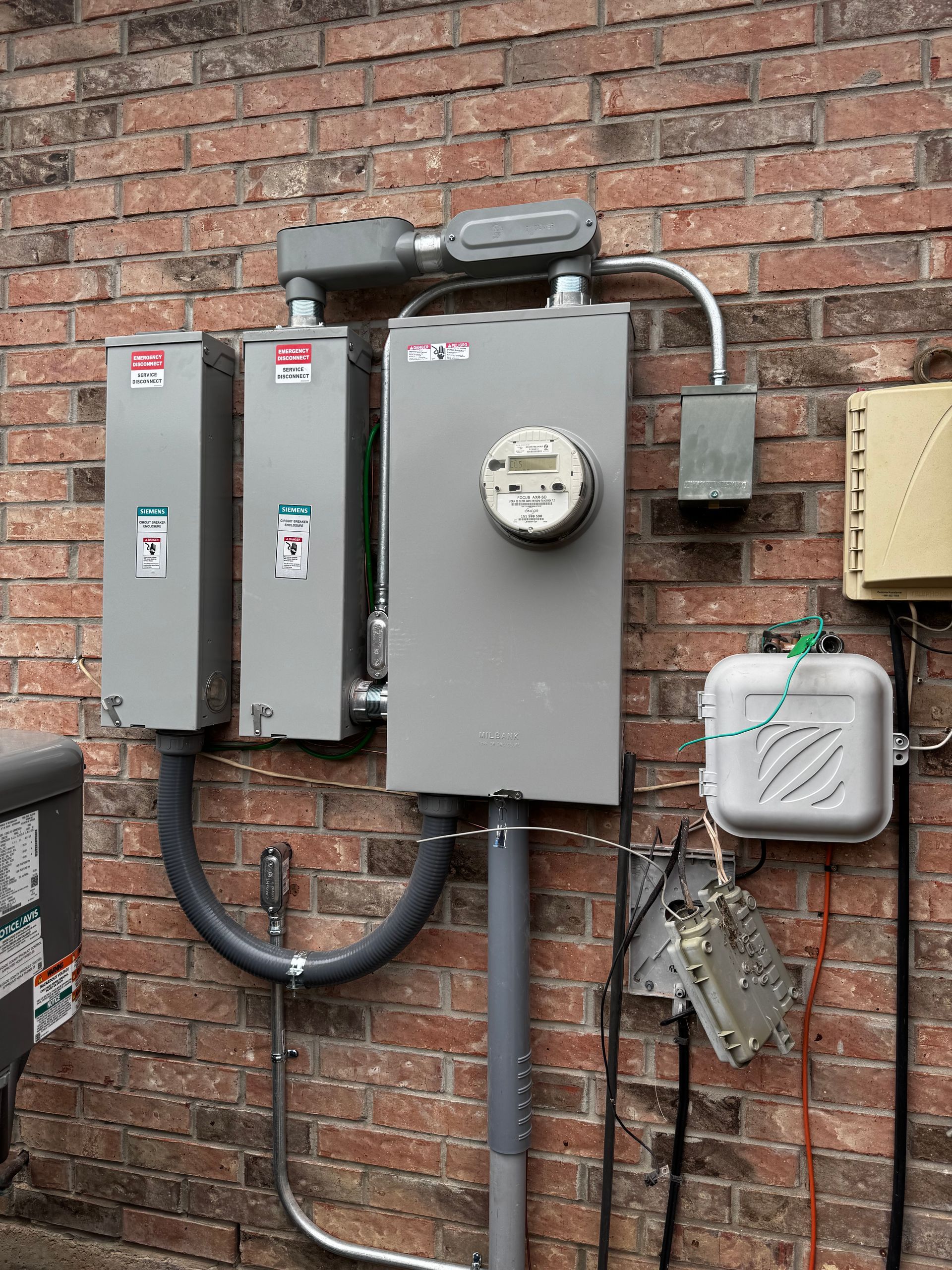 Electrical boxes and meter on a brick wall. Gray metal boxes house electrical components and a utility meter.