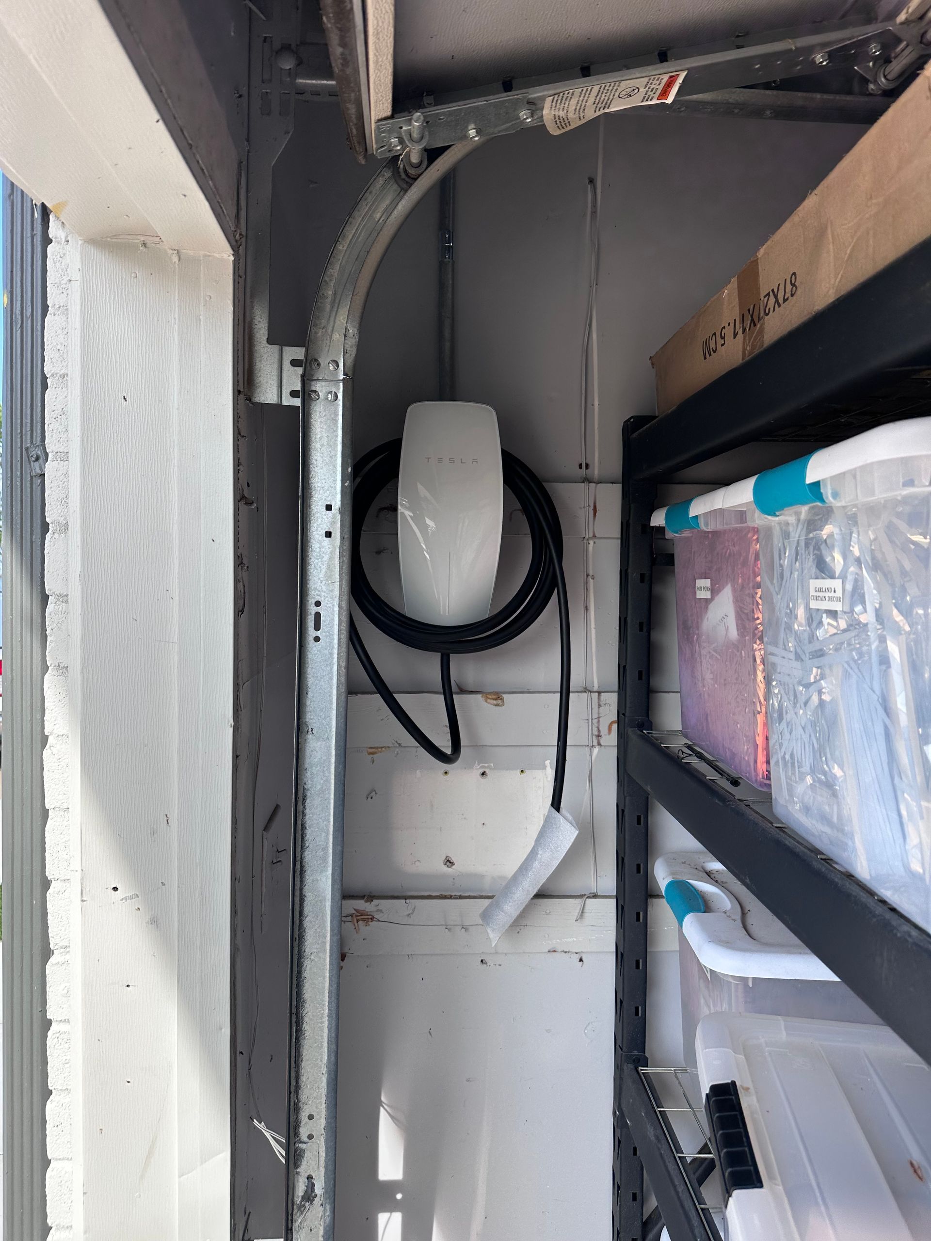 Tesla wall charger mounted on a garage wall, next to a metal shelving unit with storage bins.