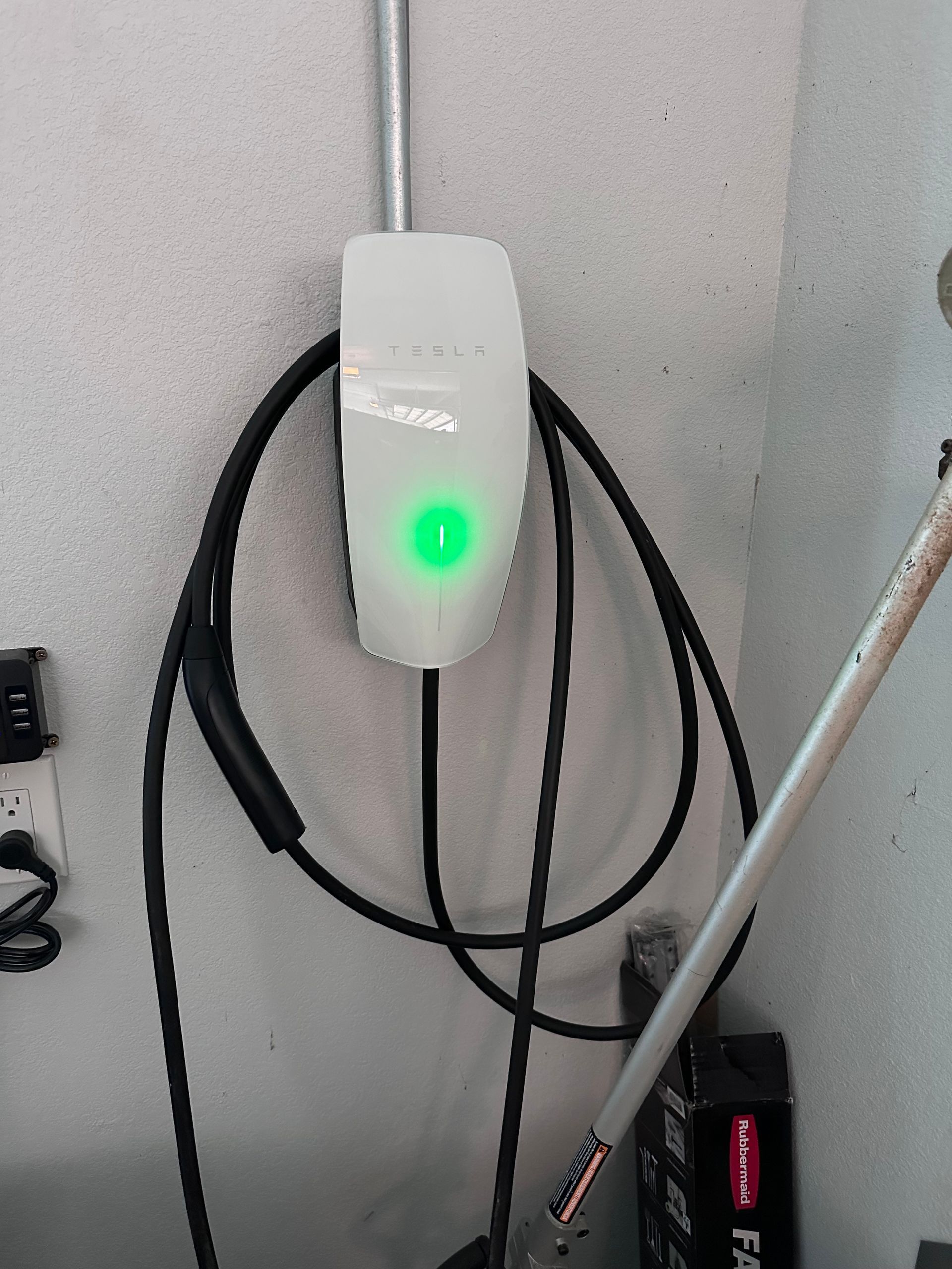White Tesla EV charger on a gray wall with a green light illuminated, cable coiled below.