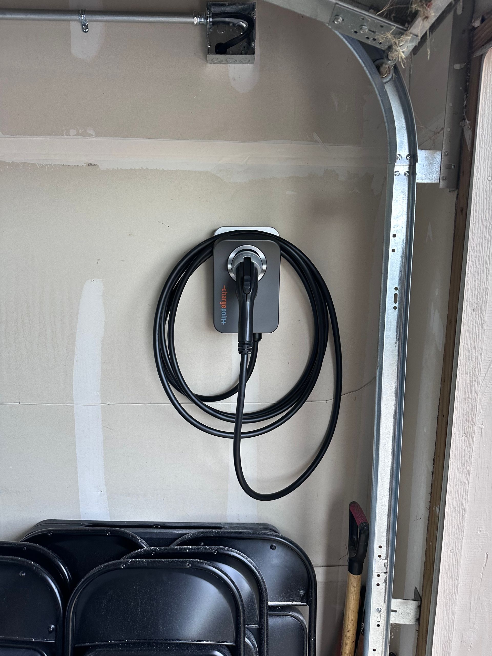 EV charger installed in a garage; gray charger, coiled black cable, black chairs, and a metal garage door frame are visible.