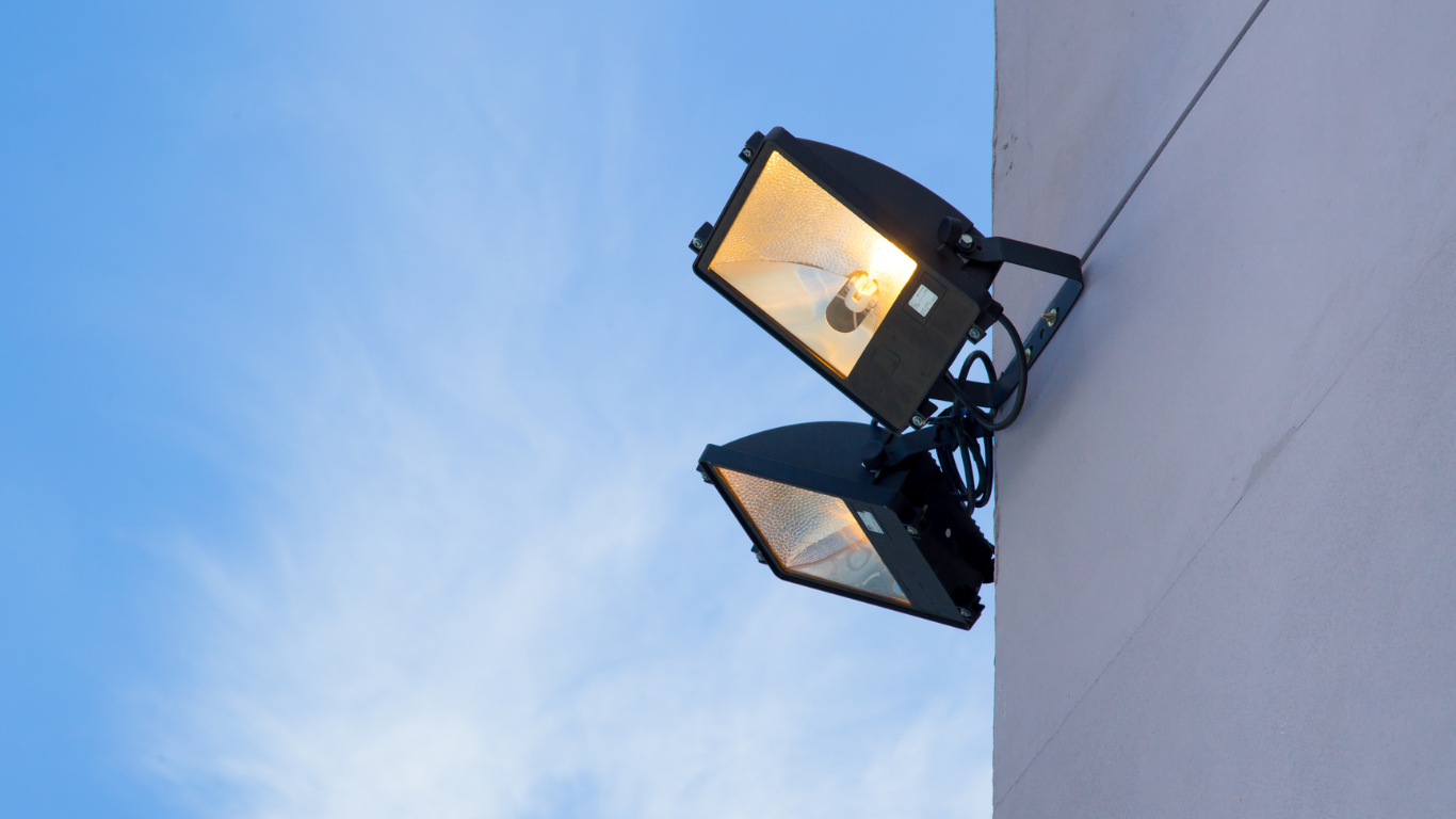 Two bright spotlights mounted on a corner of a building, illuminated against a blue sky.