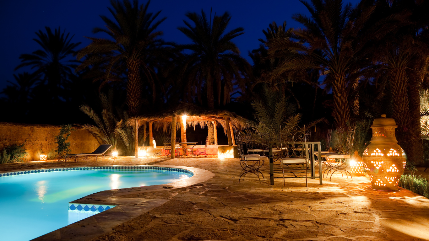 Night view of an oasis pool with a thatch-roofed cabana, lit by soft orange lights, palm trees silhouetted against a dark sky.