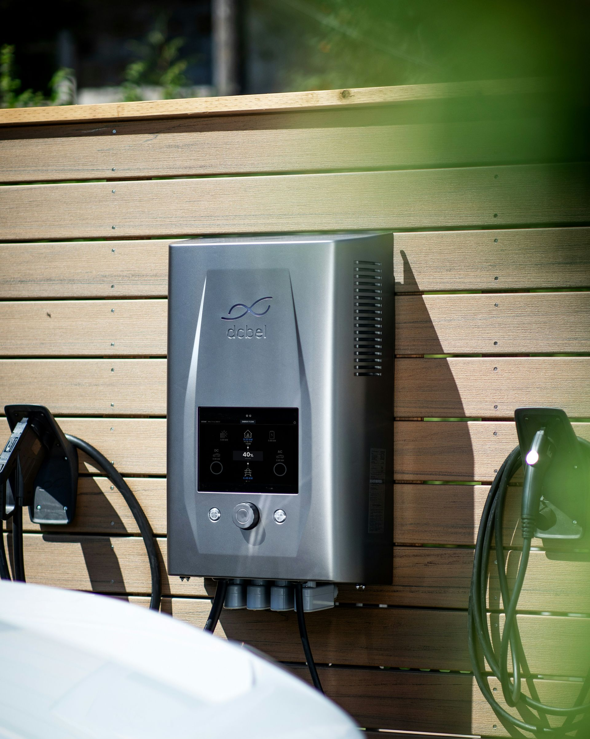 Electric vehicle charger mounted on a wooden wall, with charging cables and a car partially visible.