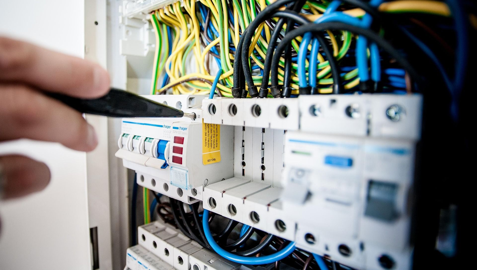Electrician's hand with tool examining wires and circuit breakers in an electrical panel; blue, yellow, and black wires.