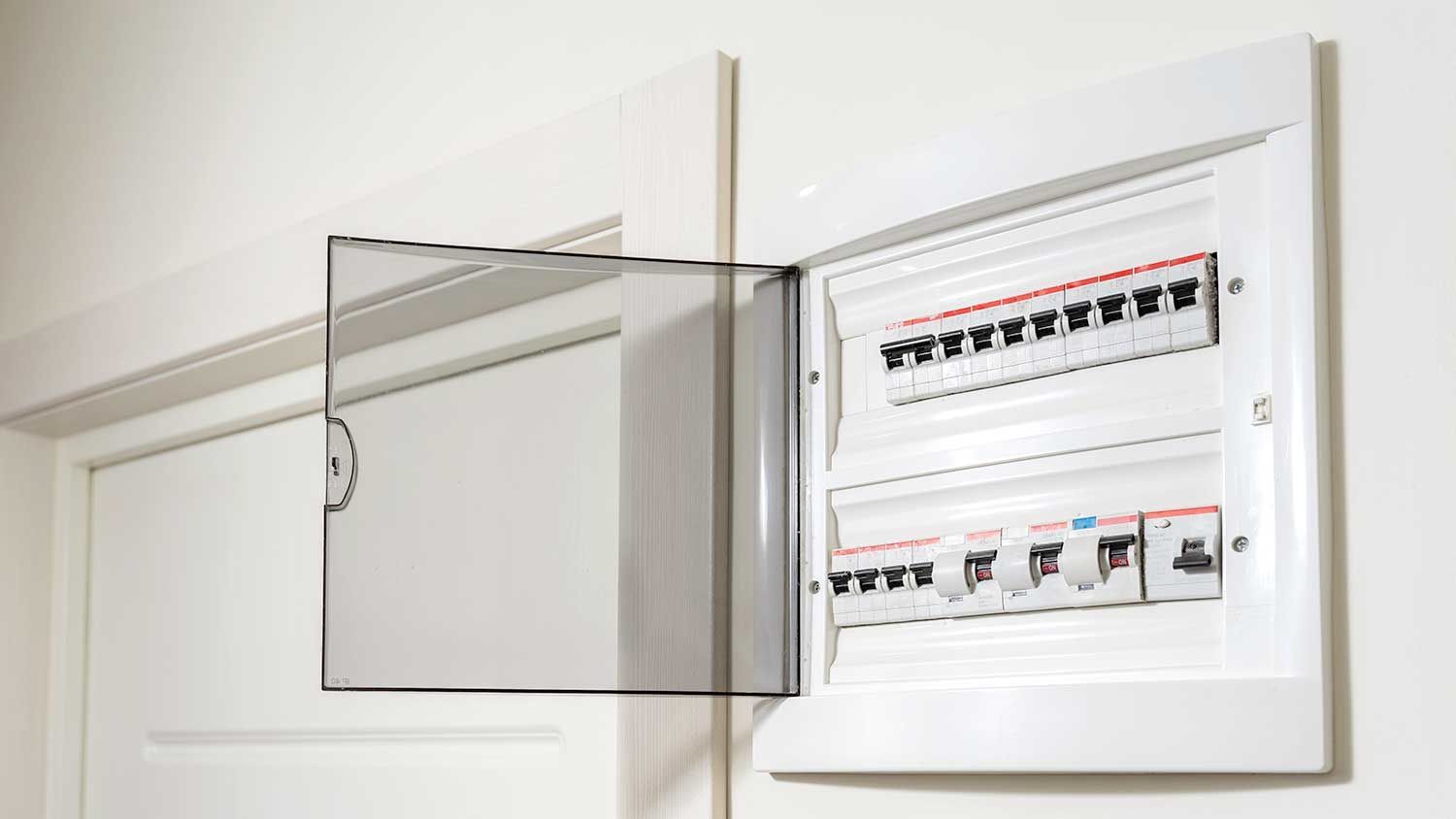White electrical panel with clear plastic door, mounted on a white wall near a door frame.
