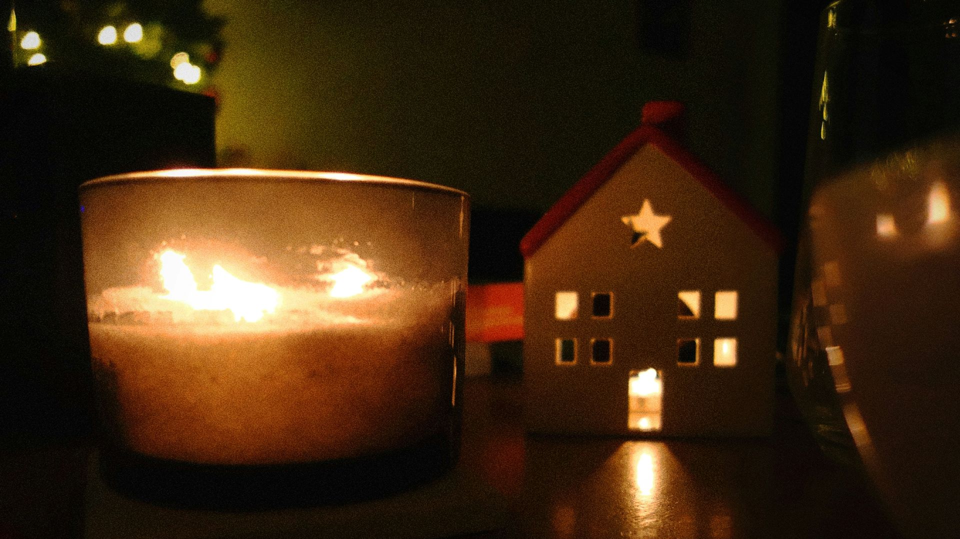 Lit candle in glass jar next to a small white house ornament with a glowing star and windows; warm, cozy scene.