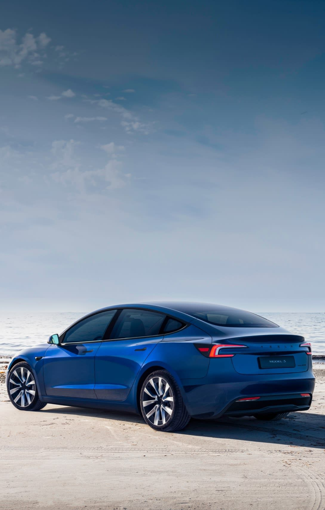 Blue Tesla sedan parked on a beach, facing the ocean under a cloudy sky.