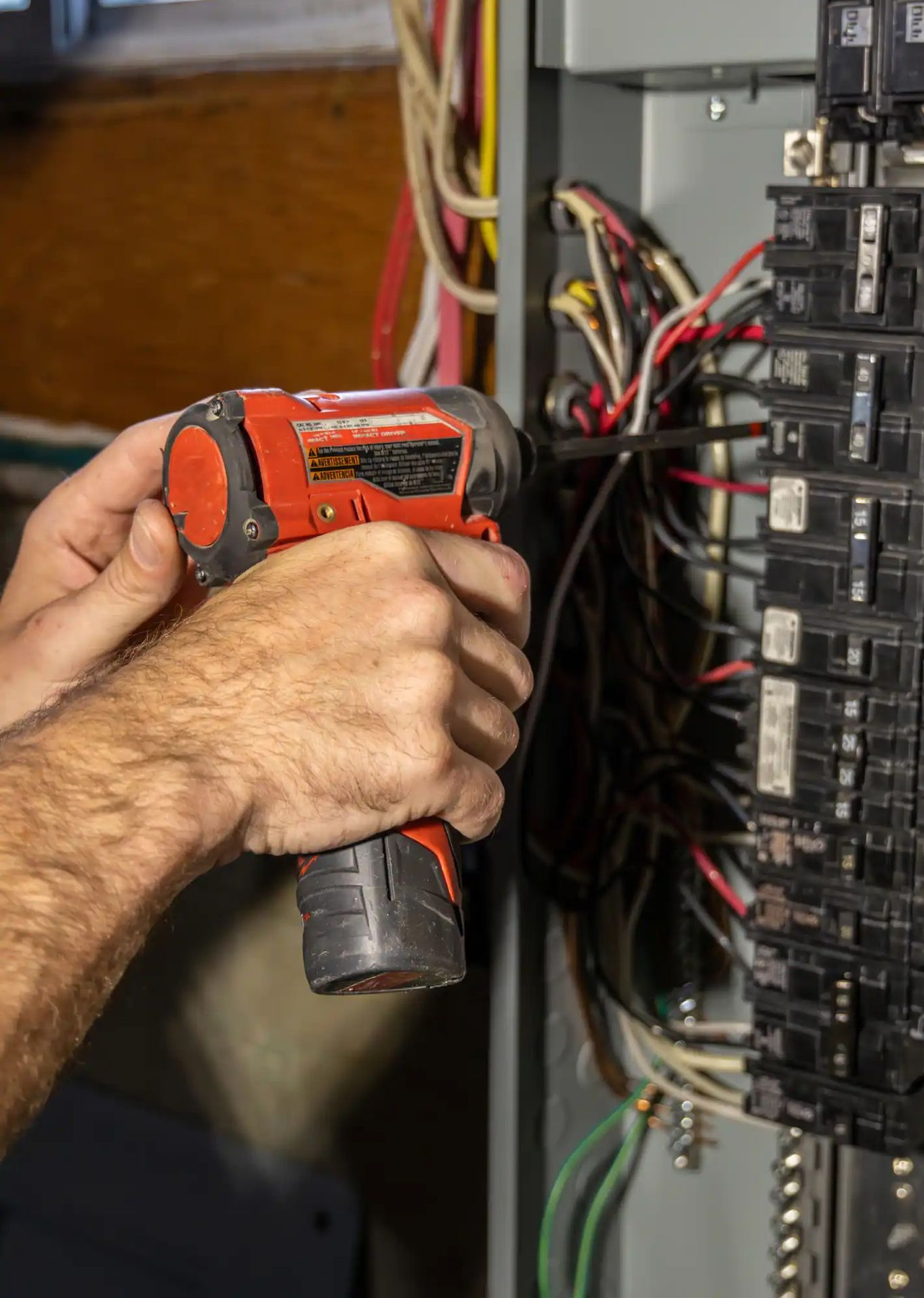 Person using a power drill to work on an electrical panel with wires and circuit breakers.