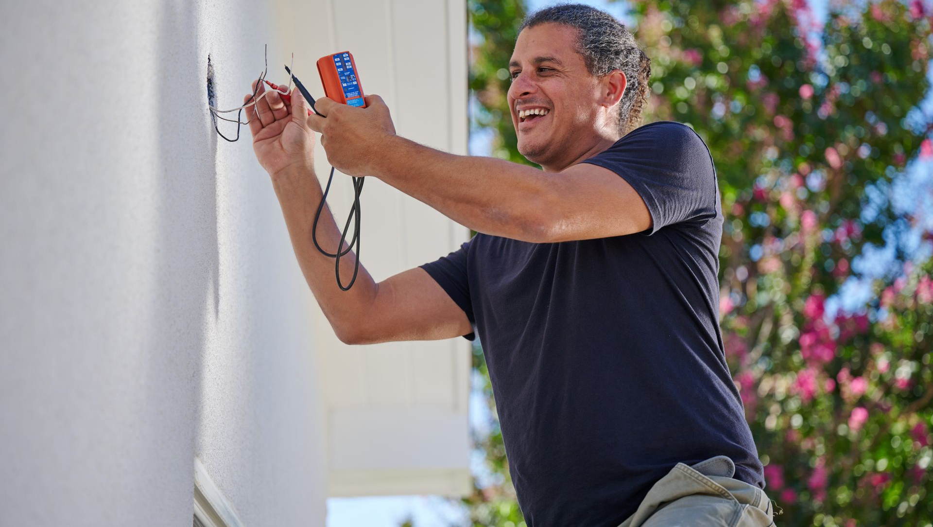 Man testing wires on a building exterior with a multimeter, smiling. Sunny day with greenery.