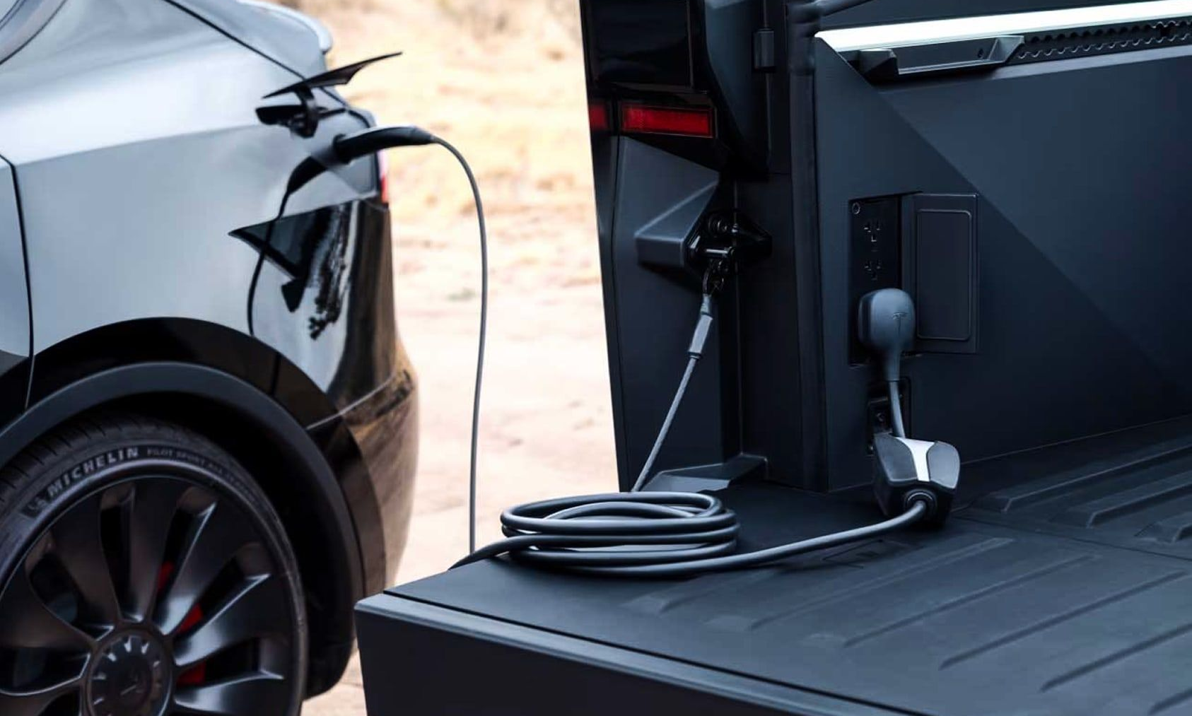 Black Tesla Model Y charging from a black truck bed outlet in an outdoor setting.
