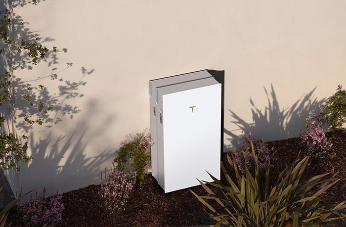 White home battery unit next to a light-colored wall, surrounded by plants and mulch.