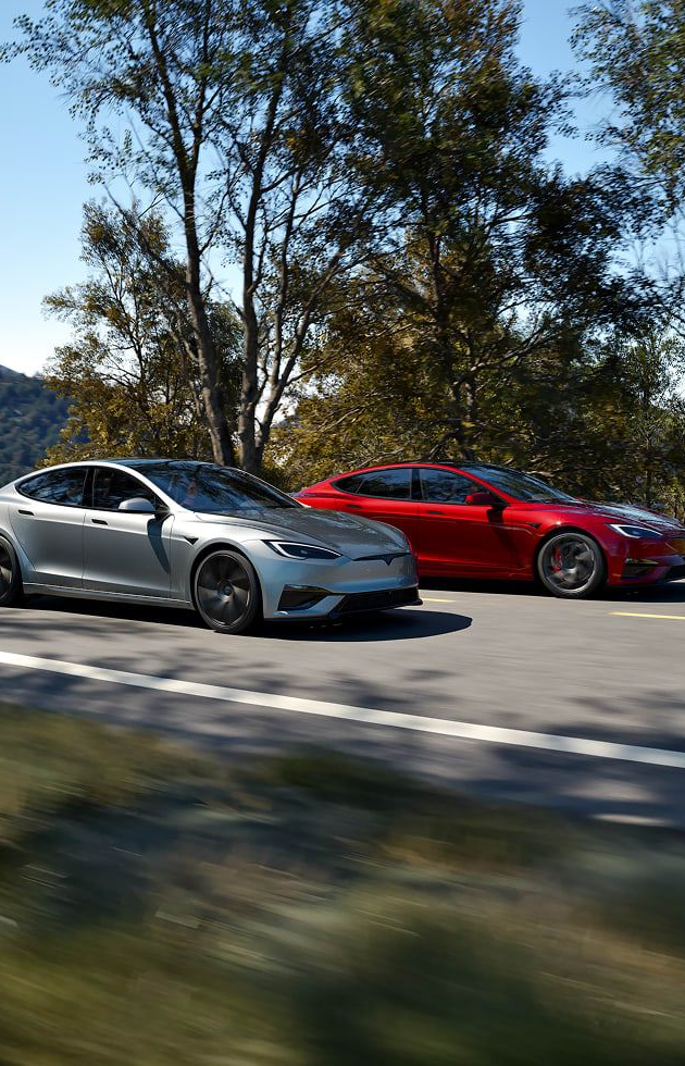 Two Tesla sedans, silver and red, driving on a road. Trees and a mountain are in the background.