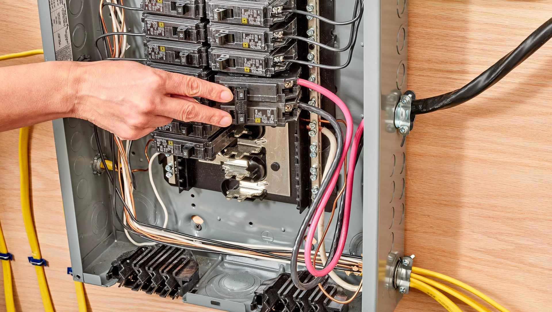 Hand pointing to a circuit breaker inside an open electrical panel, wires visible.