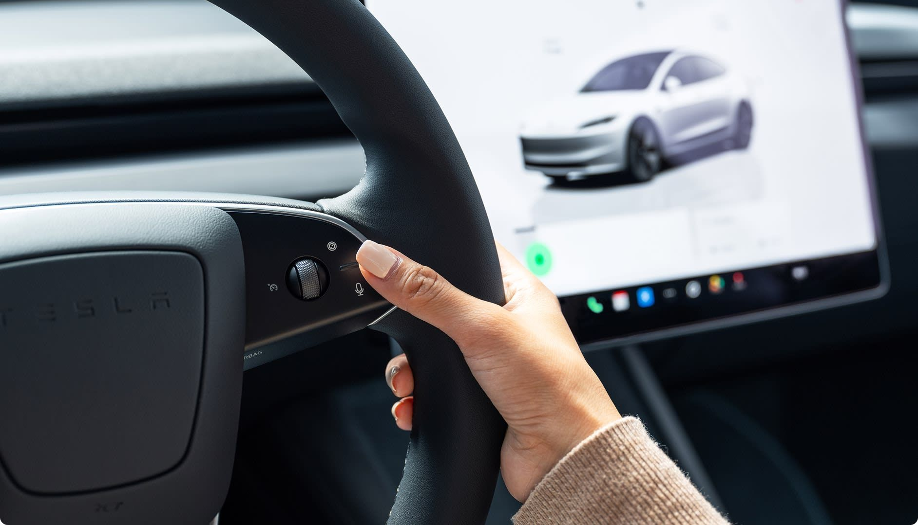 Person's hand on a black steering wheel, with a car display screen showing a silver car model in the background.