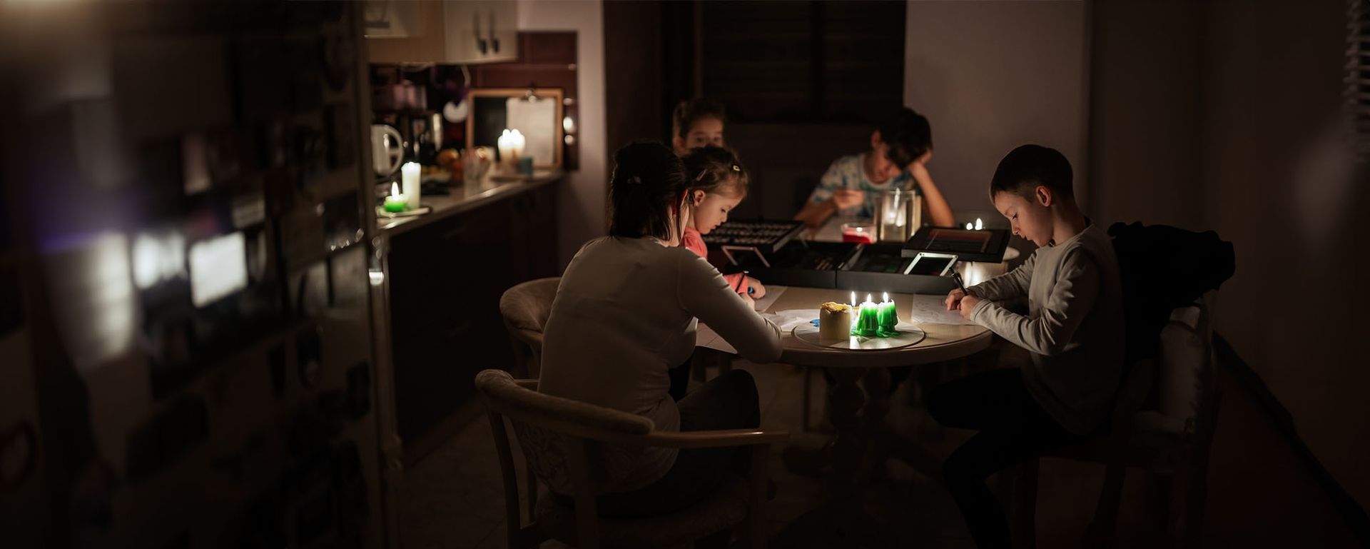 People seated around a table, writing. Dimly lit kitchen with bottles and a window.
