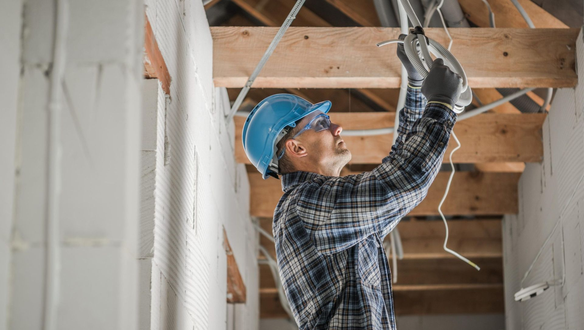 Electrician installing wires in a building's ceiling, wearing a hard hat, and safety glasses.