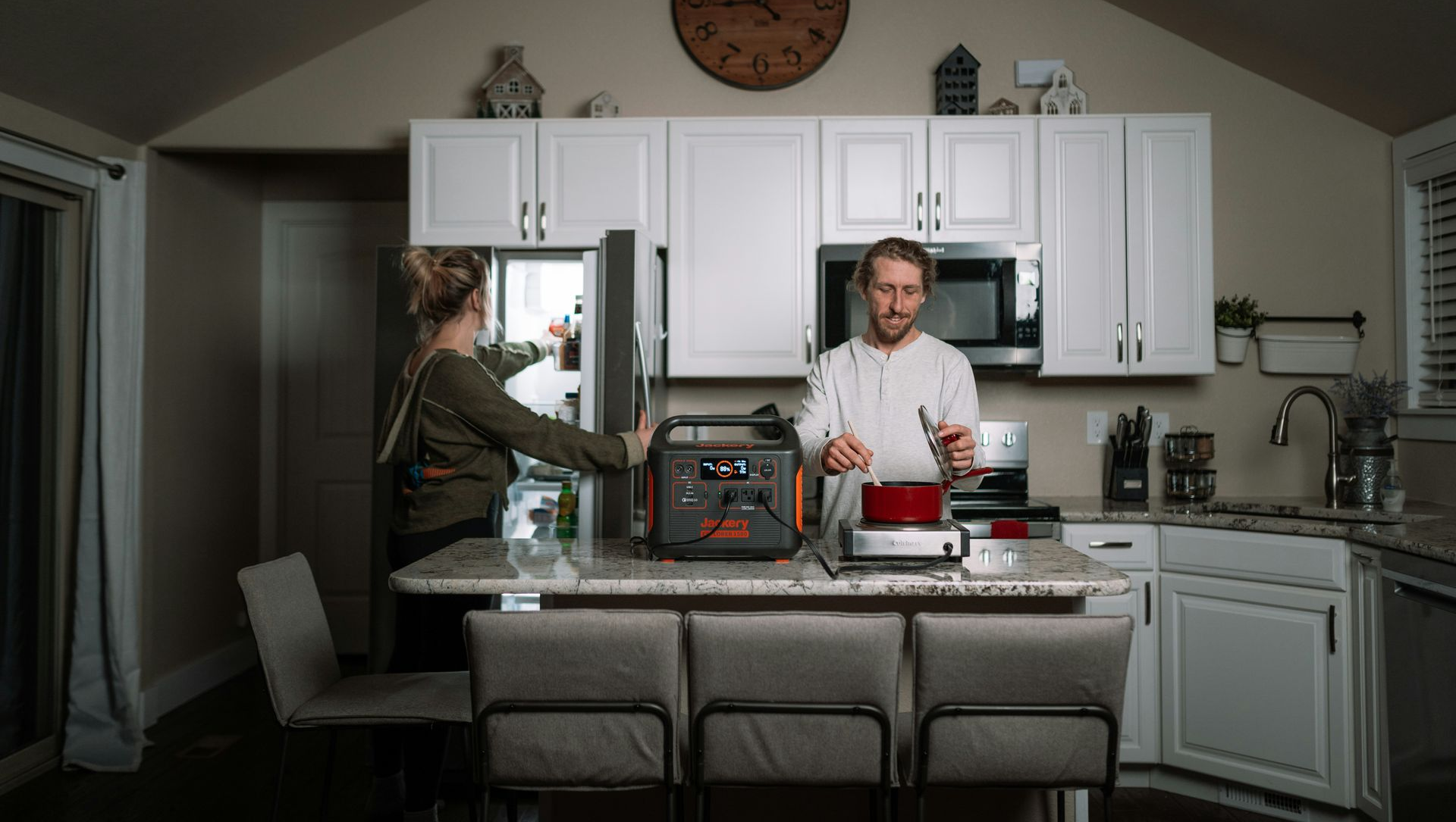 A couple in a kitchen using a generator. One opens the fridge, the other uses a cooking utensil.