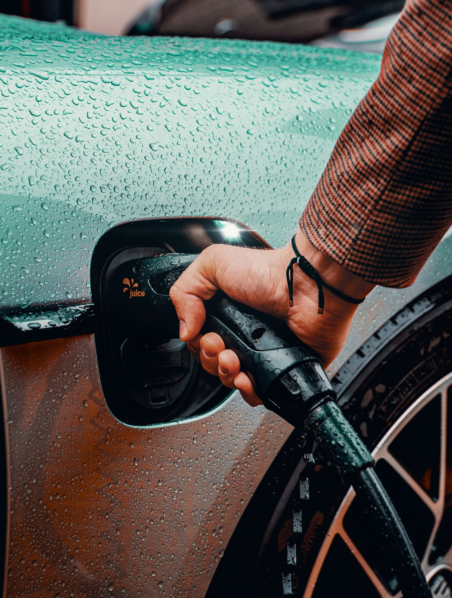 Hand plugging a charging cable into an electric car port. Raindrops on the car's silver surface.