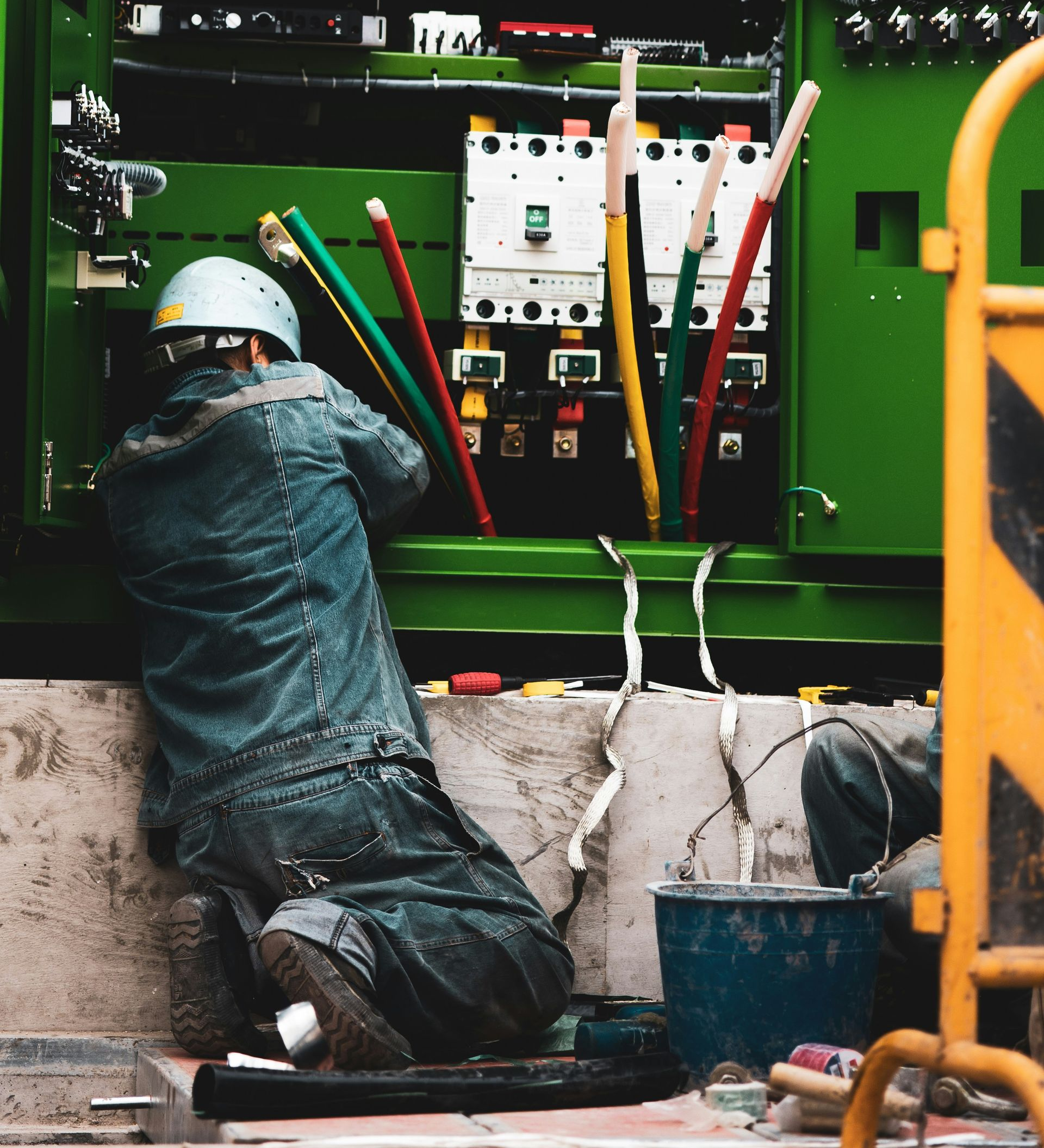 Electrician working on electrical panel, wearing safety gear; green panel, wires, outdoor setting.