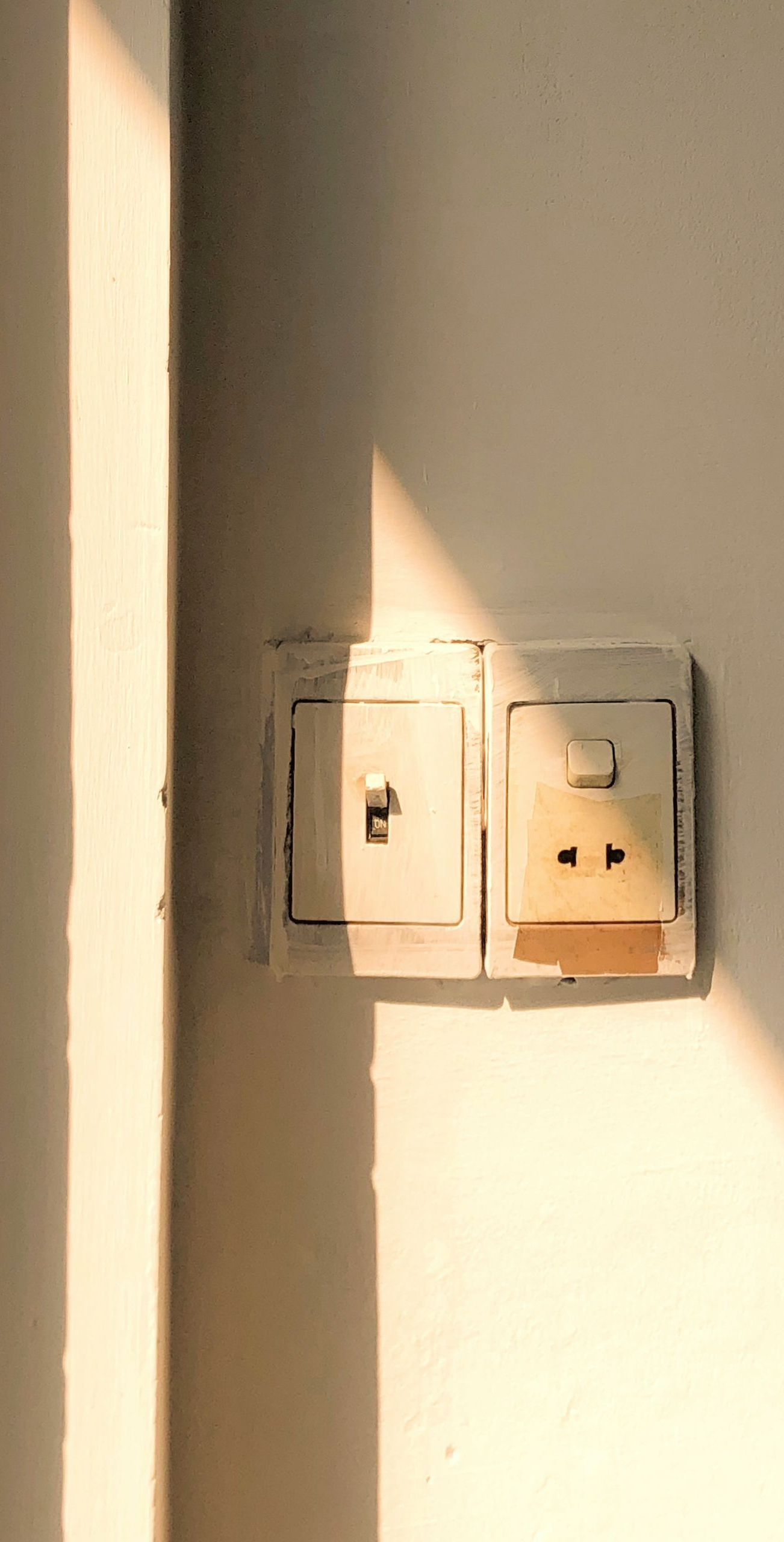 Electrical outlet and light switch on a white wall, lit by warm sunlight.