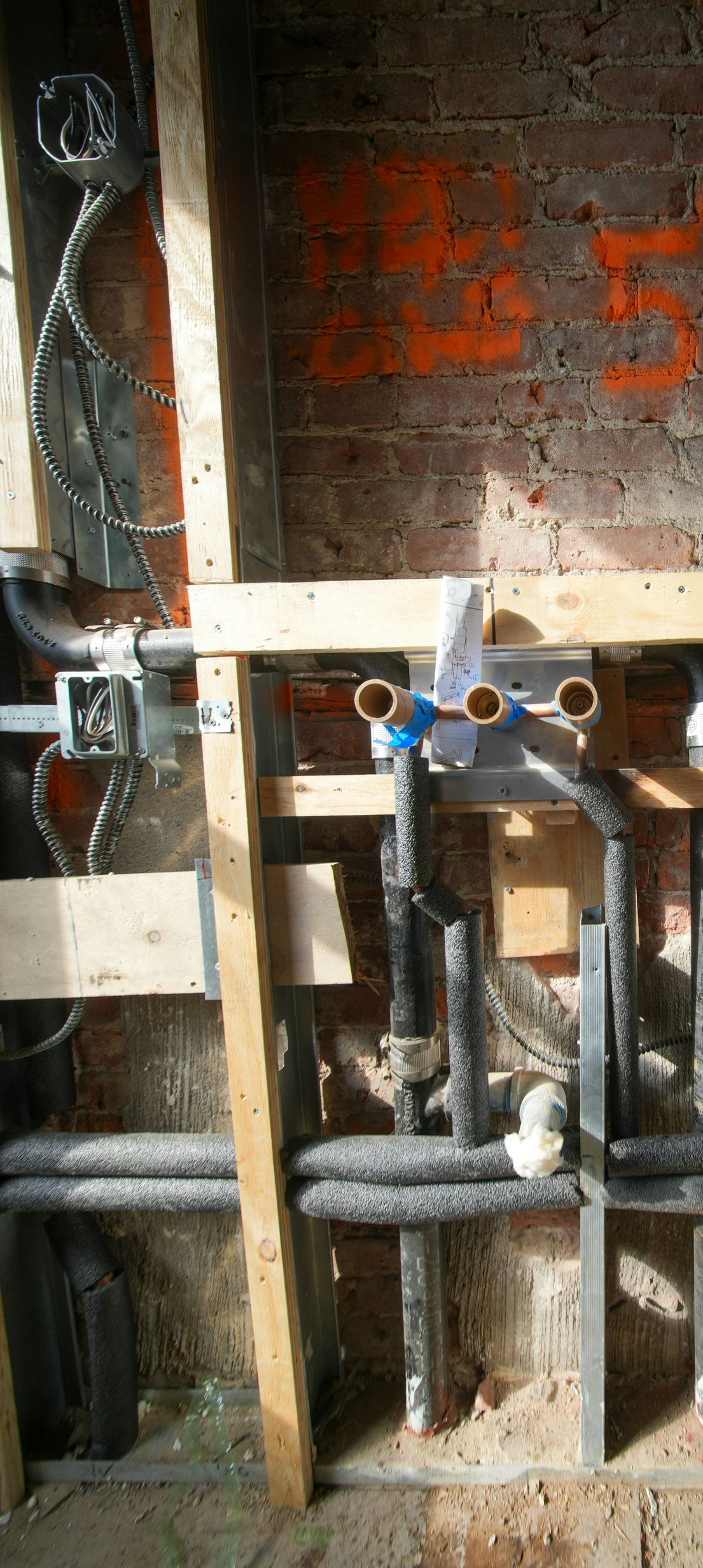 Plumbing under construction with exposed pipes and insulation within a wooden frame, against a brick wall.