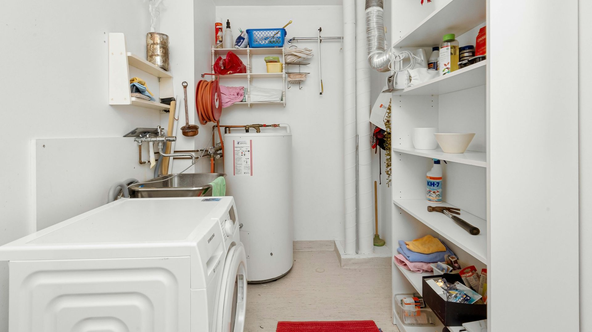 Laundry room with washing machine, water heater, shelves, and various cleaning supplies. White walls and cabinetry.