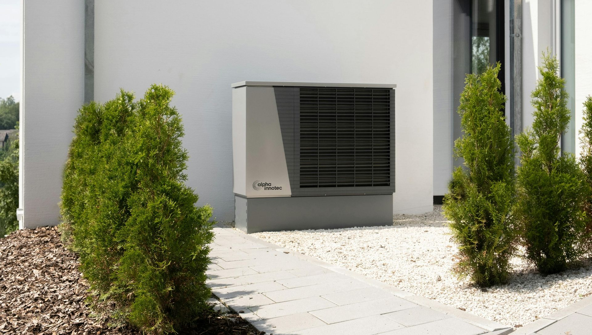 Heat pump unit next to a building, surrounded by green shrubs and white stones.