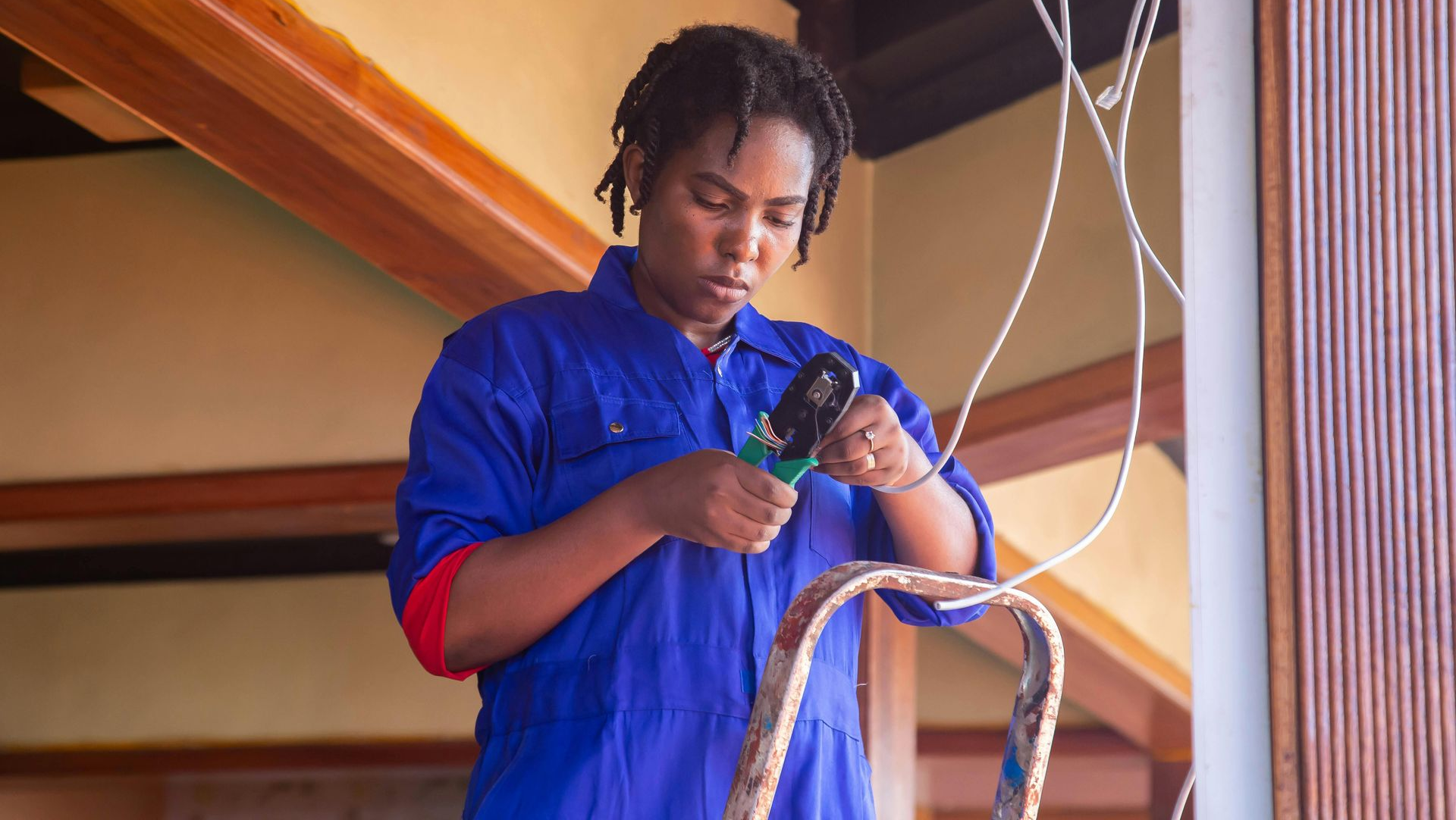 Woman in blue overalls on a ladder, using pliers to work with electrical wires indoors.