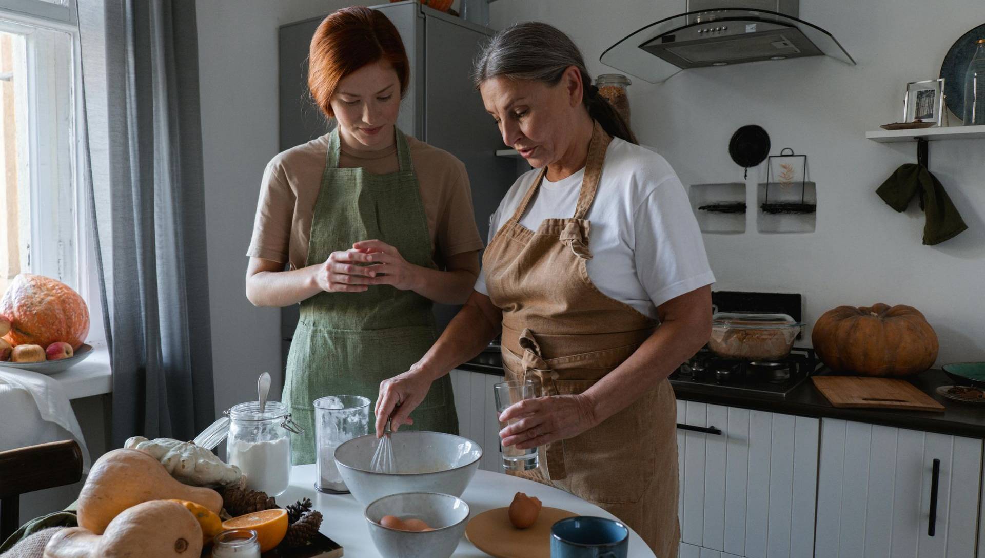 Two people in aprons baking together in a kitchen, one whisking ingredients, the other watching.