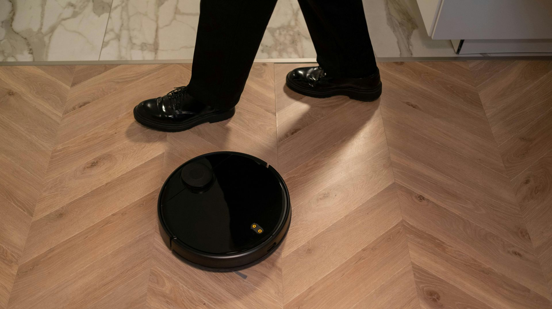Black robotic vacuum cleaner on wooden floor with a person's legs in view.