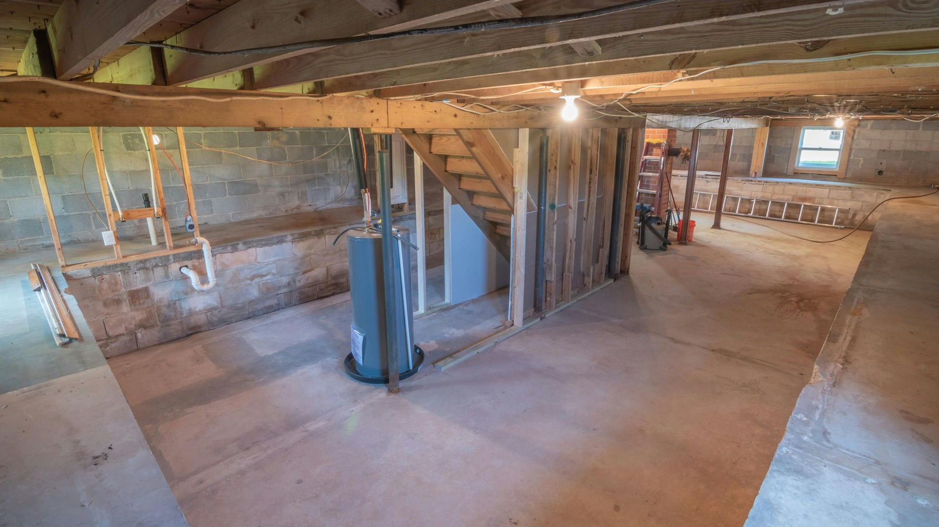 Basement with exposed beams, concrete floor, water heater, and stairs leading upwards.