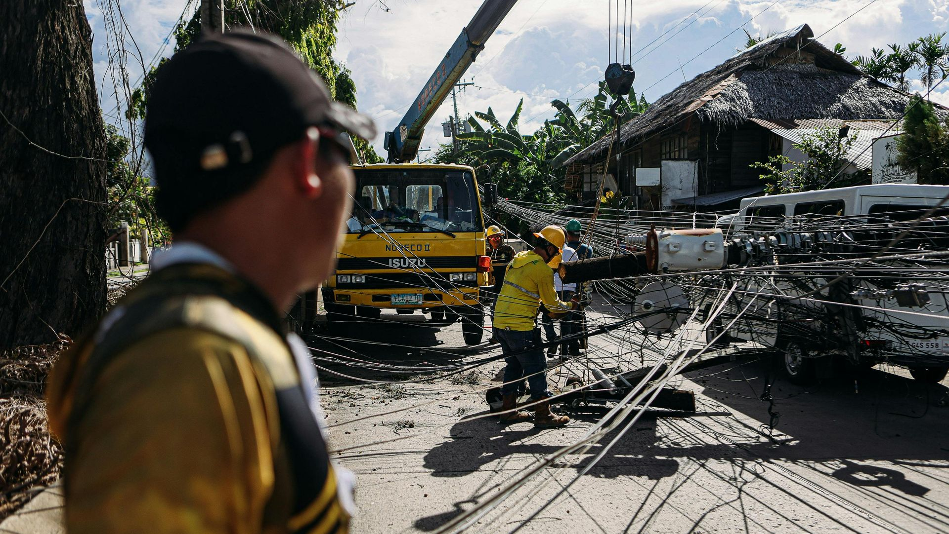 Man watches workers clearing debris with a crane and truck in a damaged street.