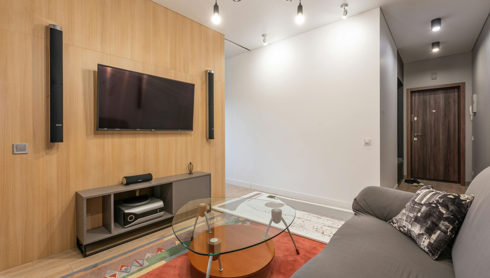 Living room with TV, speakers, and sofa. Wooden paneling, red rug, and glass coffee table.