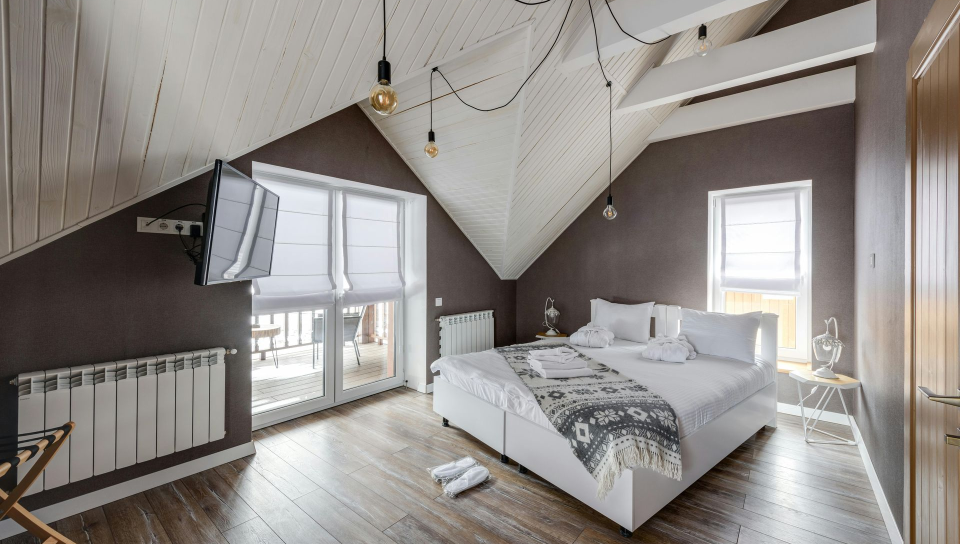 Bedroom in a slanted-roof attic: white bed, dark walls, window, radiator, exposed beams, and hanging lights.