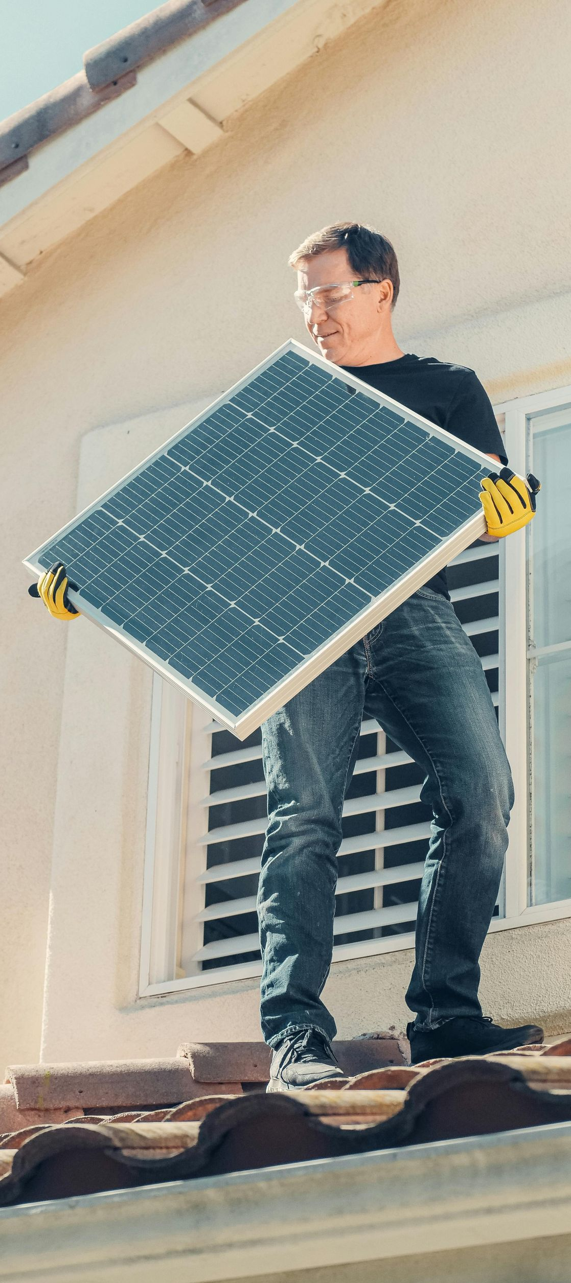 Man in jeans and black shirt carrying a solar panel on a roof.