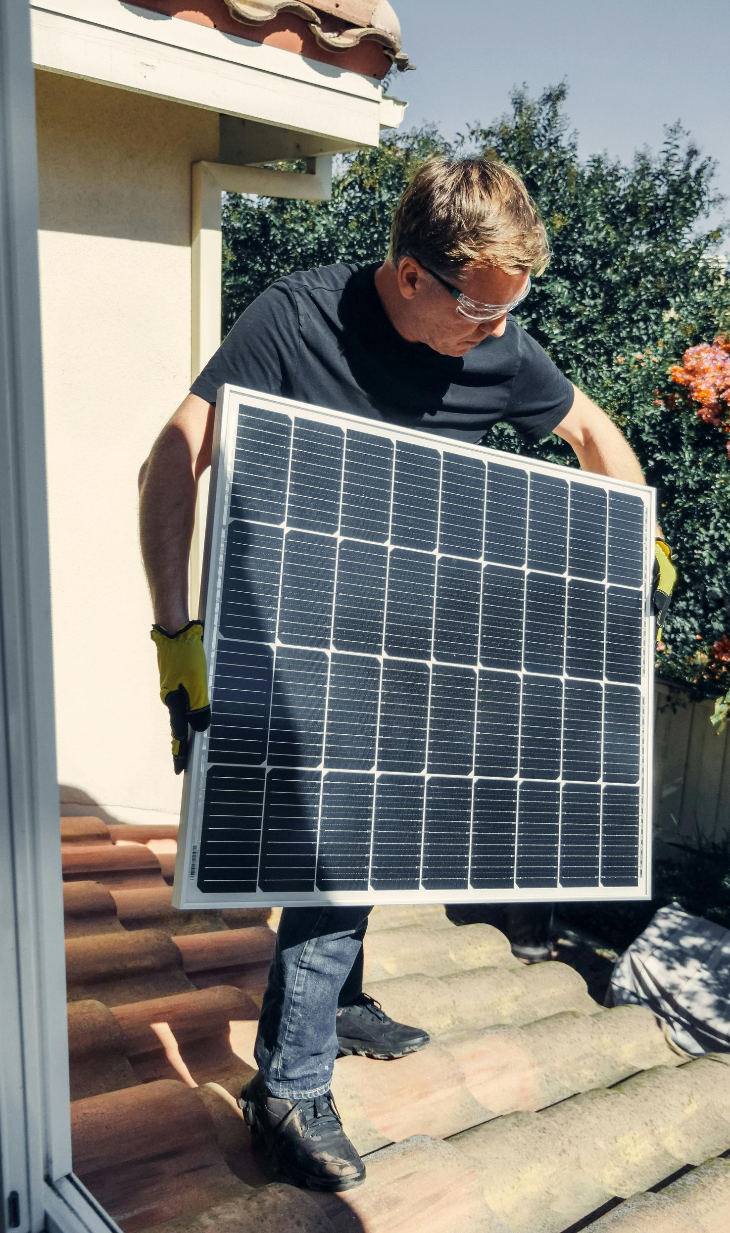 A person in a black shirt carries a solar panel on a tiled roof.