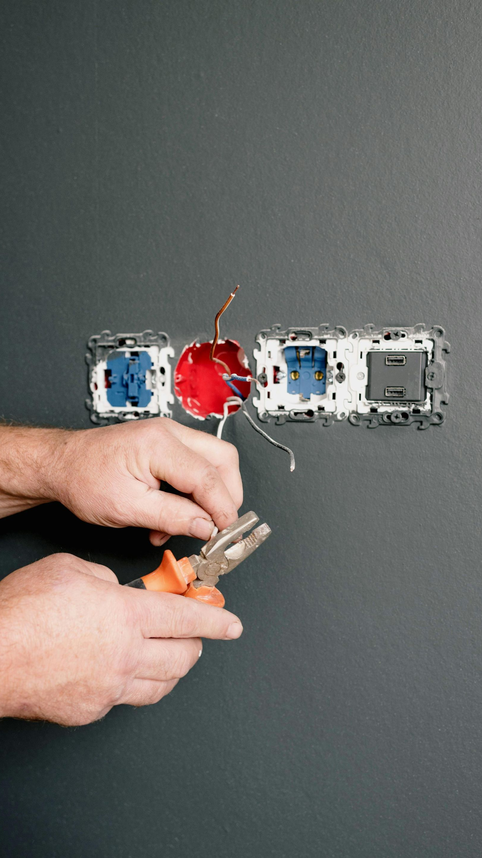 Hands using pliers to work on electrical wiring in a wall outlet box.
