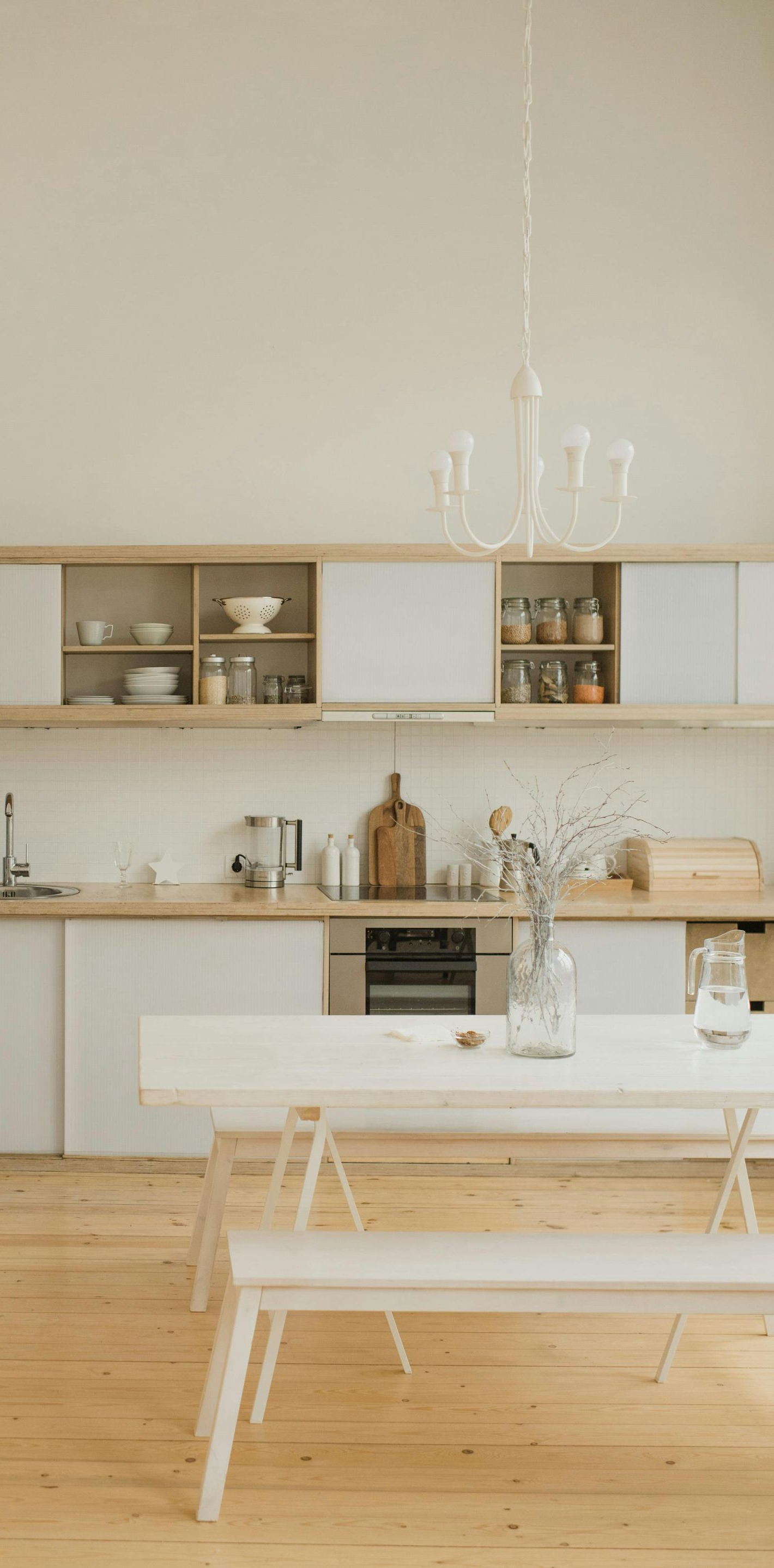 Light-filled kitchen with a long white table, benches, and pale wood floors. Cabinets and a chandelier.