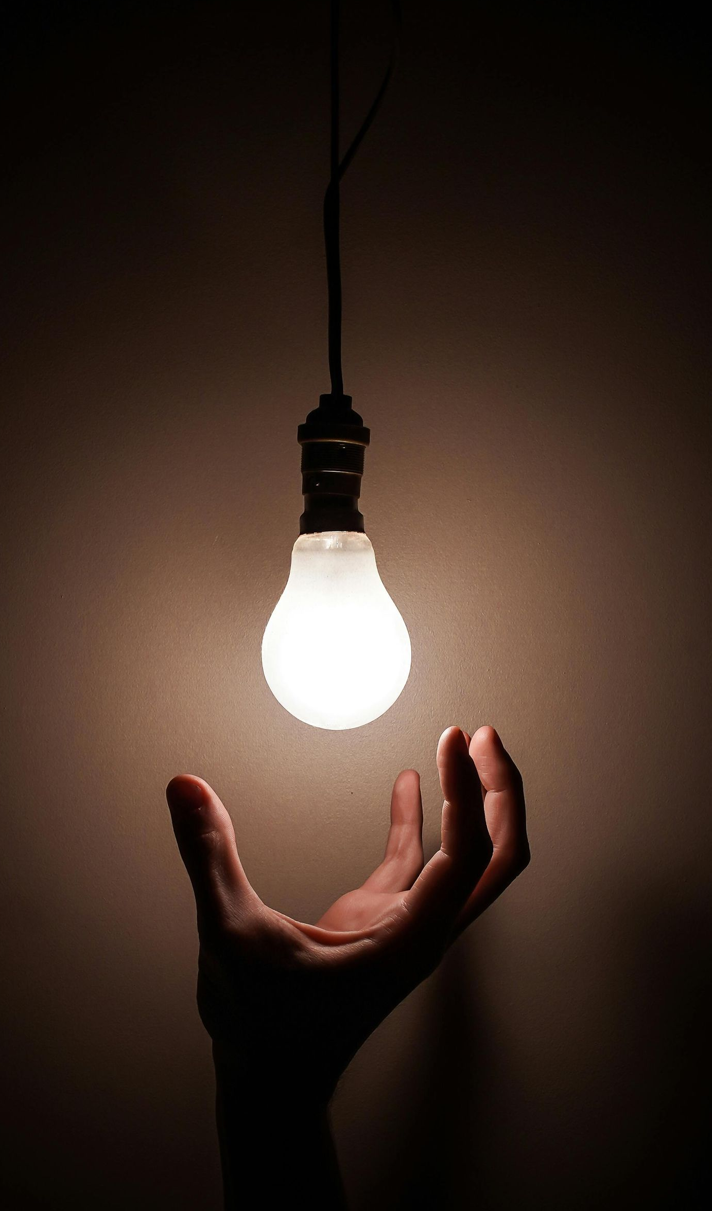 Hand reaching up toward a glowing lightbulb hanging from a wire in darkness.
