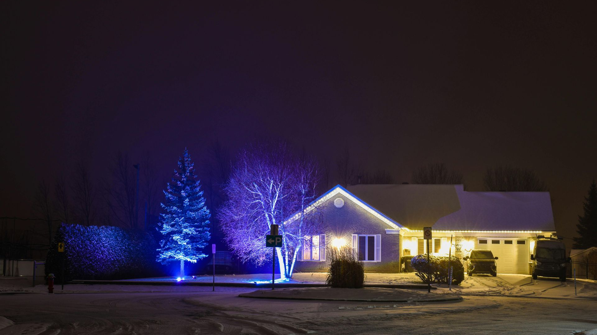 House and trees with blue and white Christmas lights, covered in snow at night.
