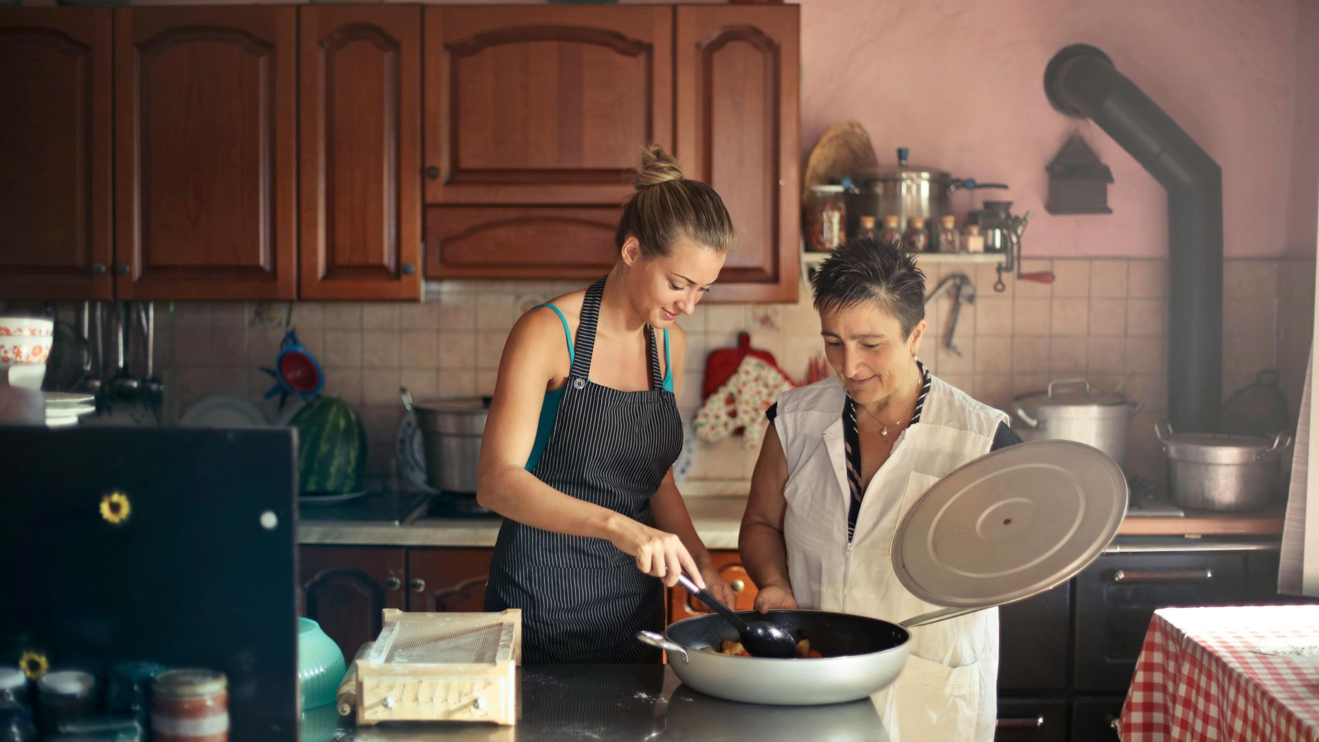 Woman in apron stirring food with another woman in a kitchen with wooden cabinets and a stove.