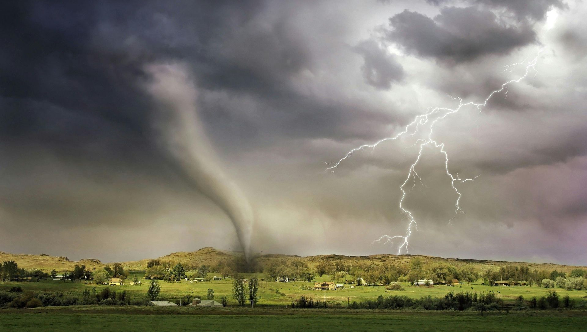 Tornado with dark funnel descends over green landscape; lightning strikes amidst stormy clouds.