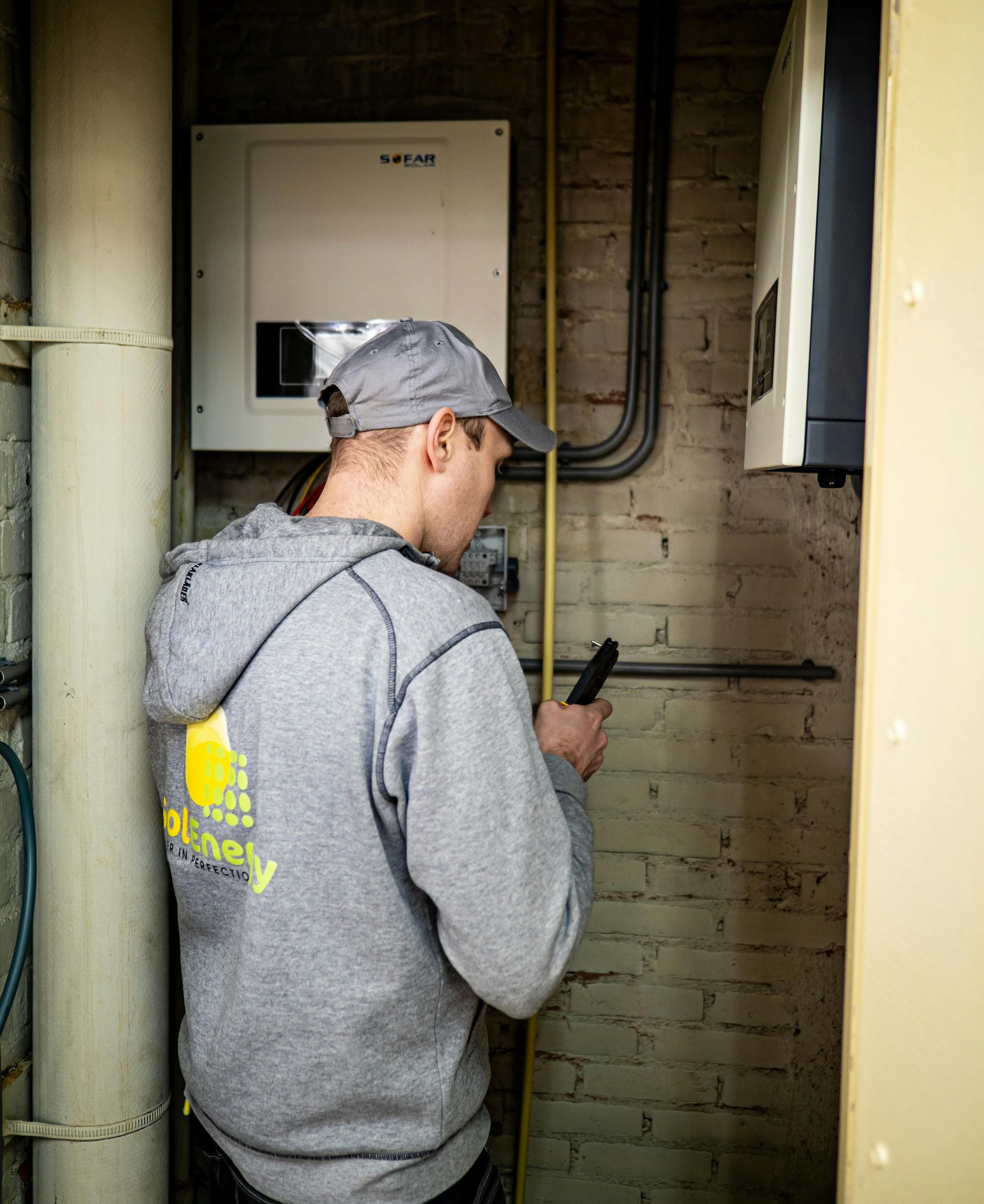 A person in a gray hoodie and hat examines electrical equipment in a brick-walled room.