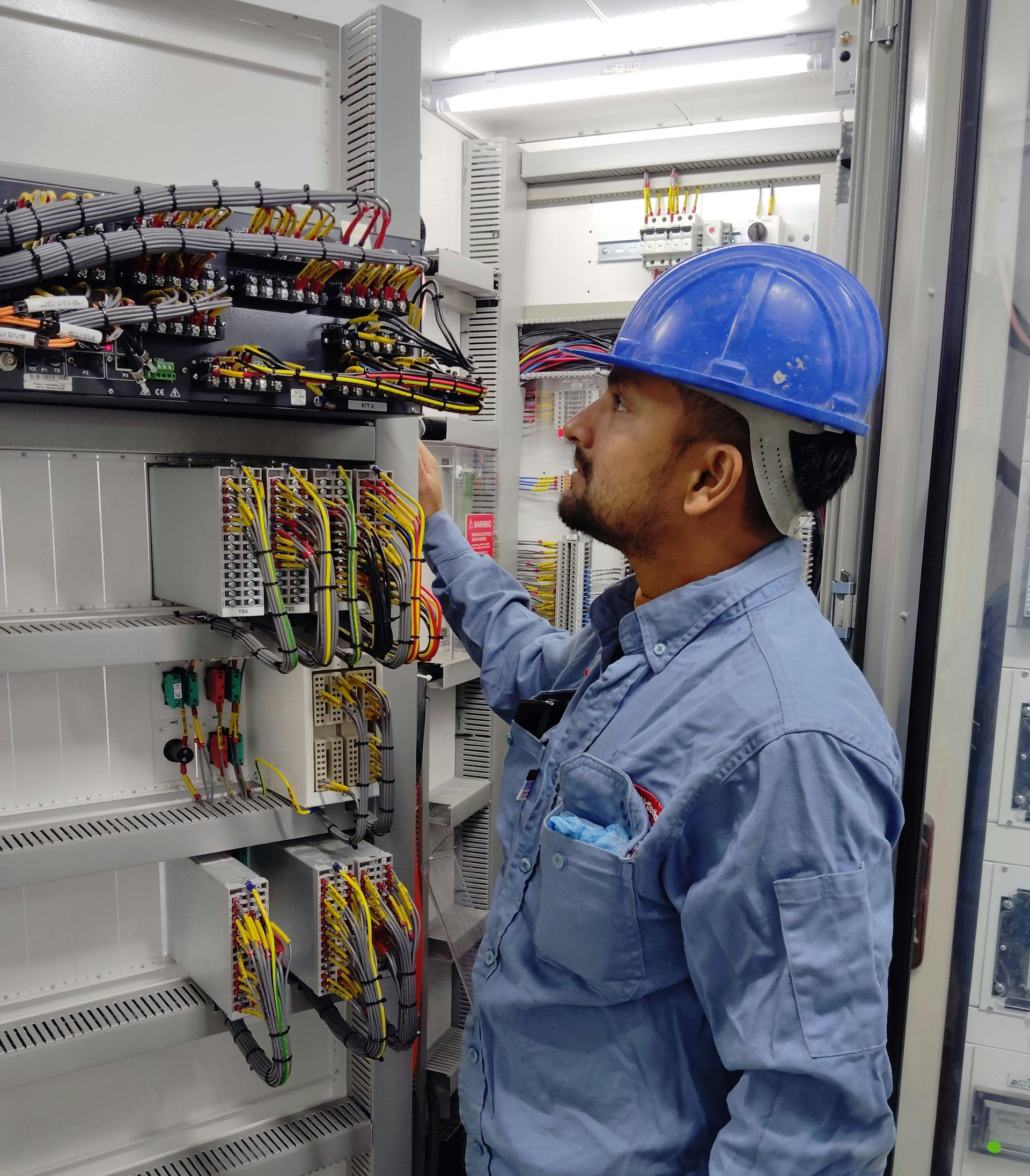 Man in blue hard hat inspects electrical panel wiring.