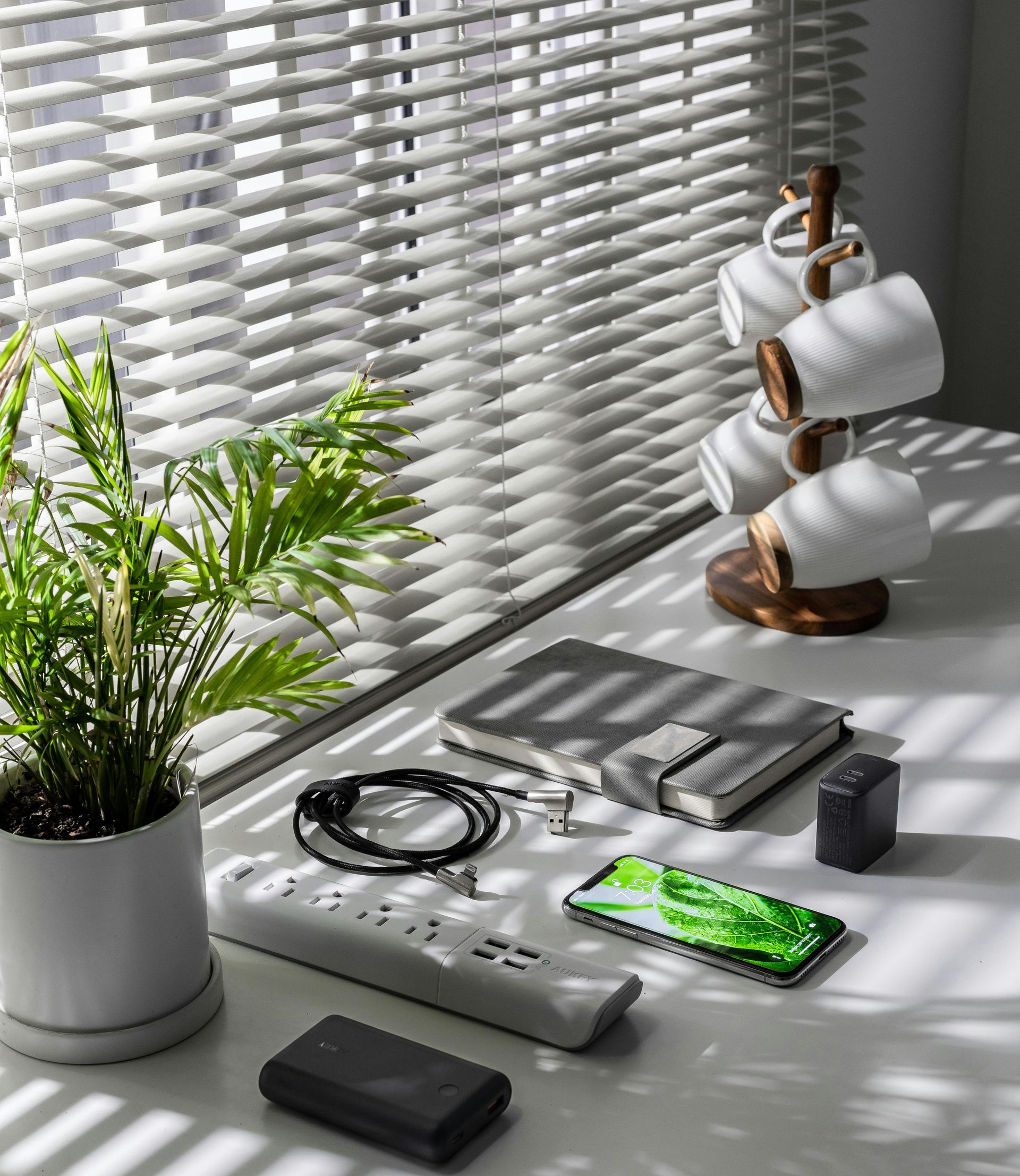 Desk scene: plant, coffee mugs, electronics (phone, power strip, charger), notebook, blinds.