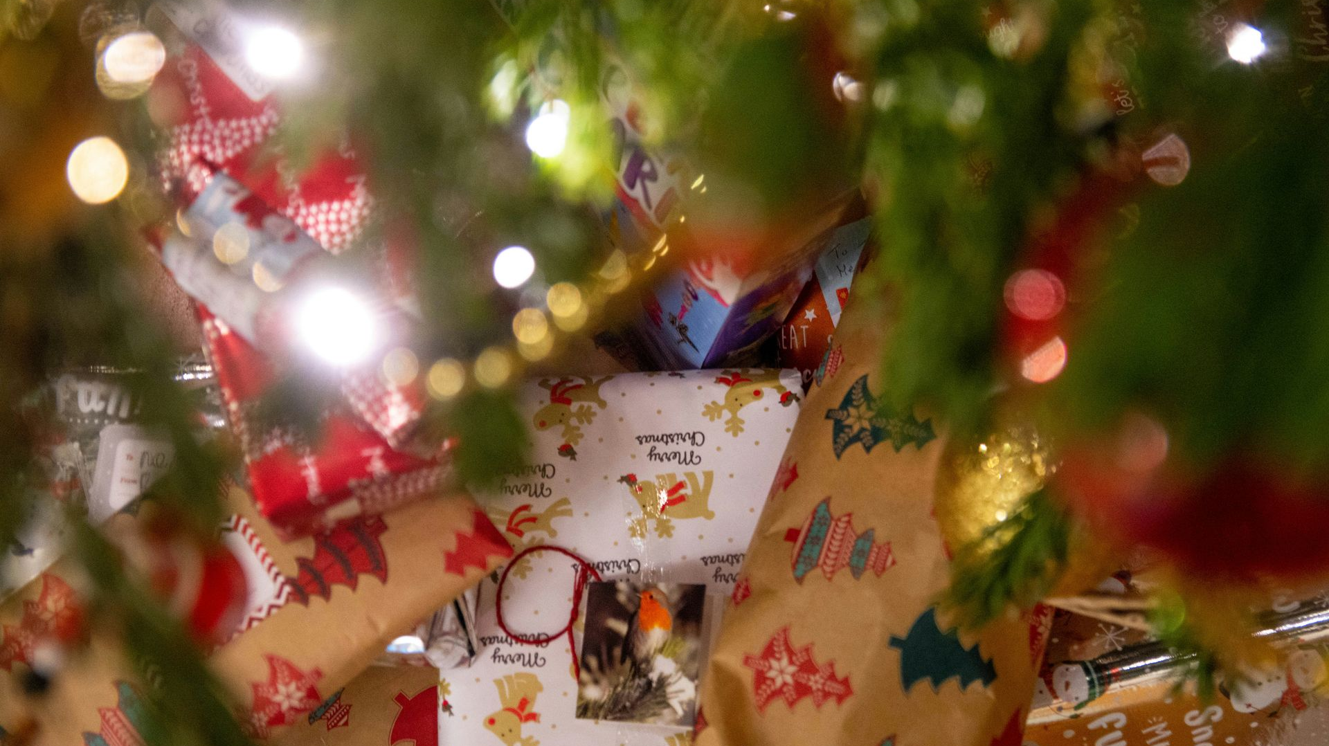 Christmas gifts wrapped in festive paper under a decorated tree, with string lights.