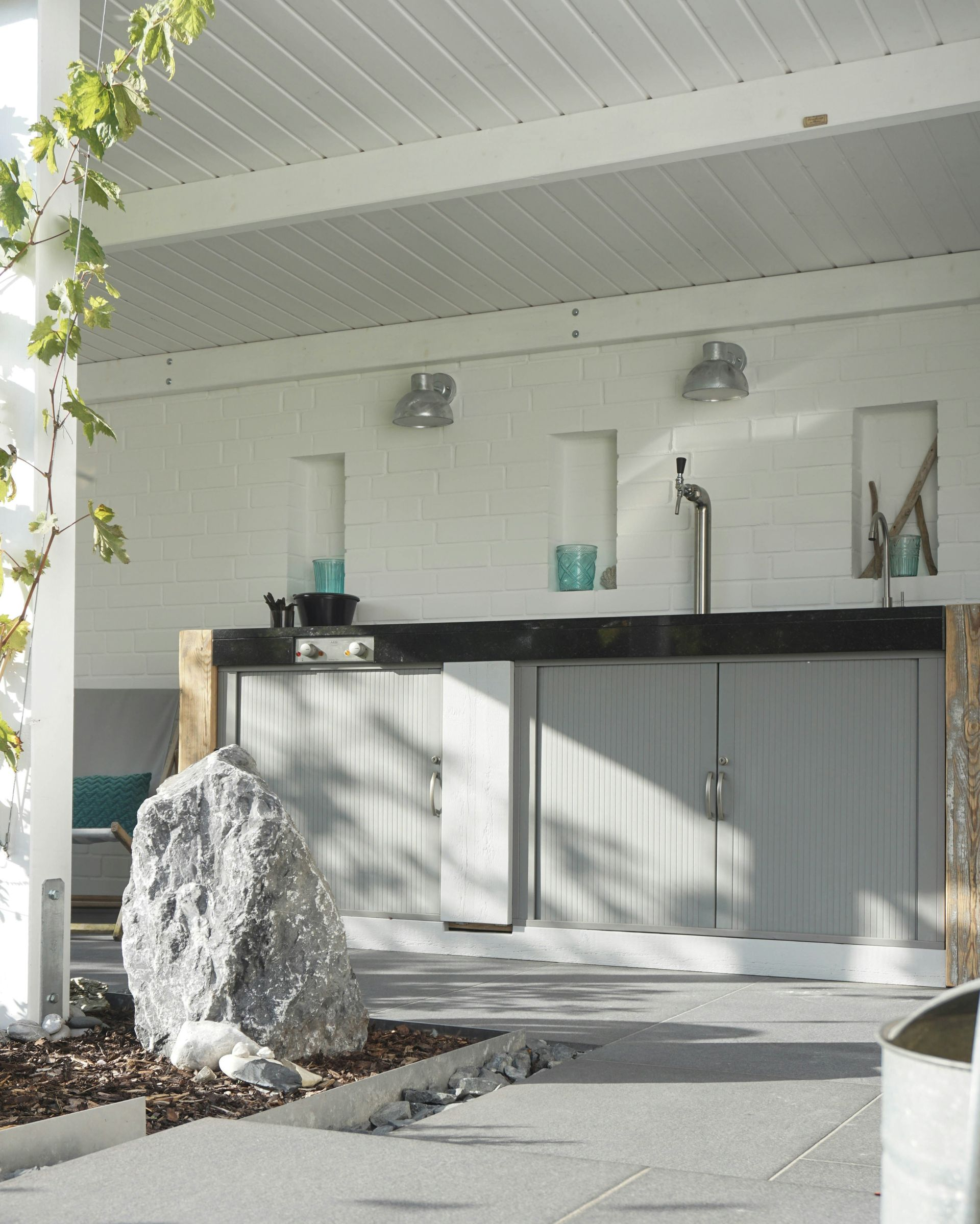 Outdoor bar area with white walls, gray cabinets, a large rock, and overhead lighting.