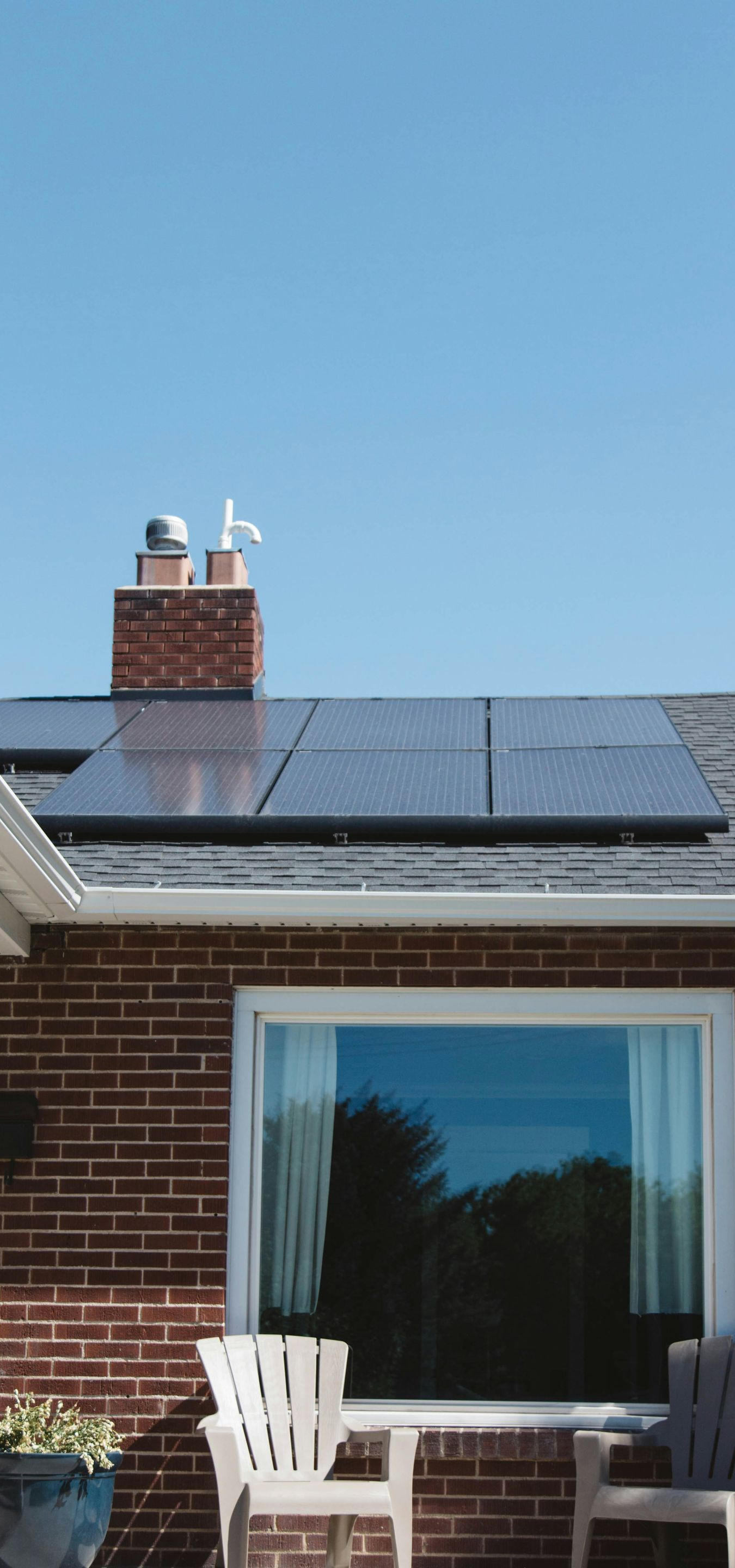 Solar panels on a brick house roof with a chimney and window, under a clear blue sky.