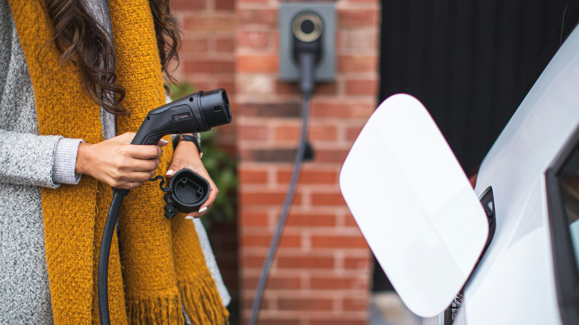 Woman holding EV charging cable near a white car and a home charging station.