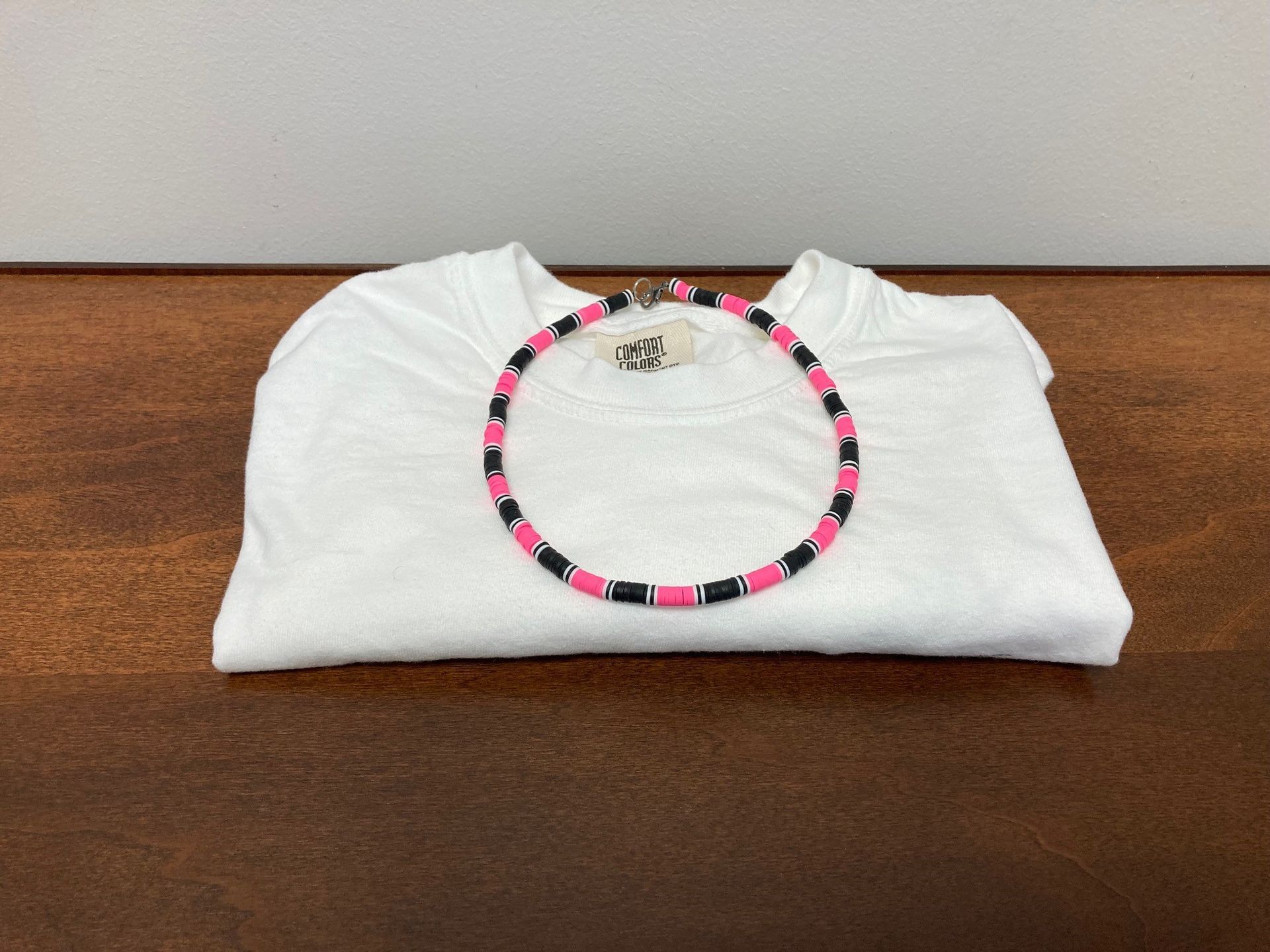 White folded t-shirt with a pink and black beaded necklace on a wooden surface.
